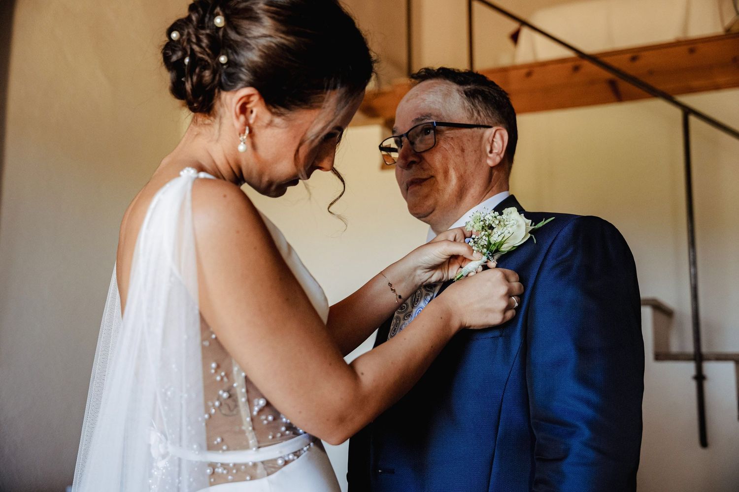 Bride in a sheer wedding gown pinning a boutonniere on an older man in a navy suit.