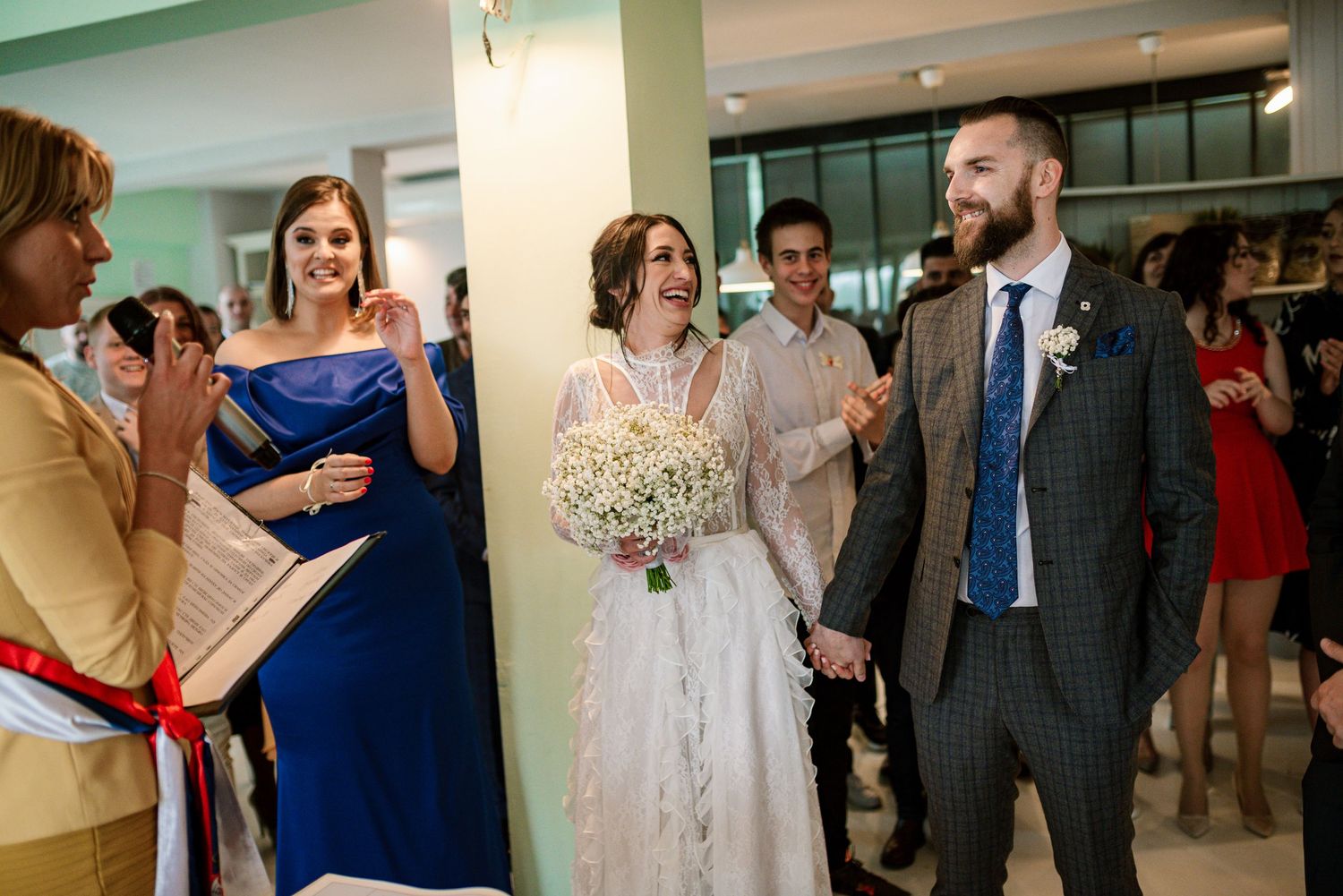 Joyful bride holding bouquet and groom in checkered suit celebrating at indoor wedding ceremony.