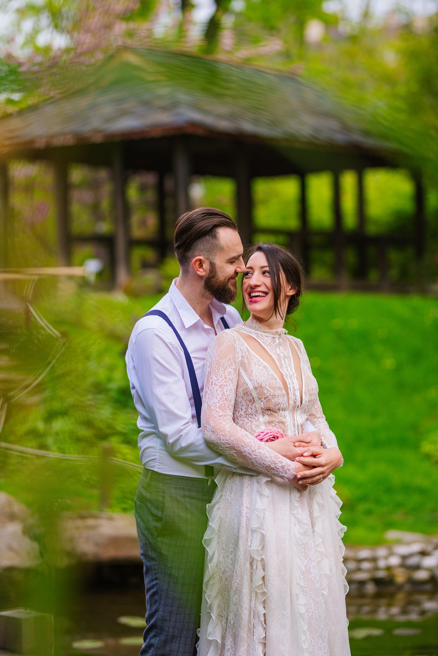 Groom embracing laughing bride in front of a wooden gazebo in a scenic green garden setting.