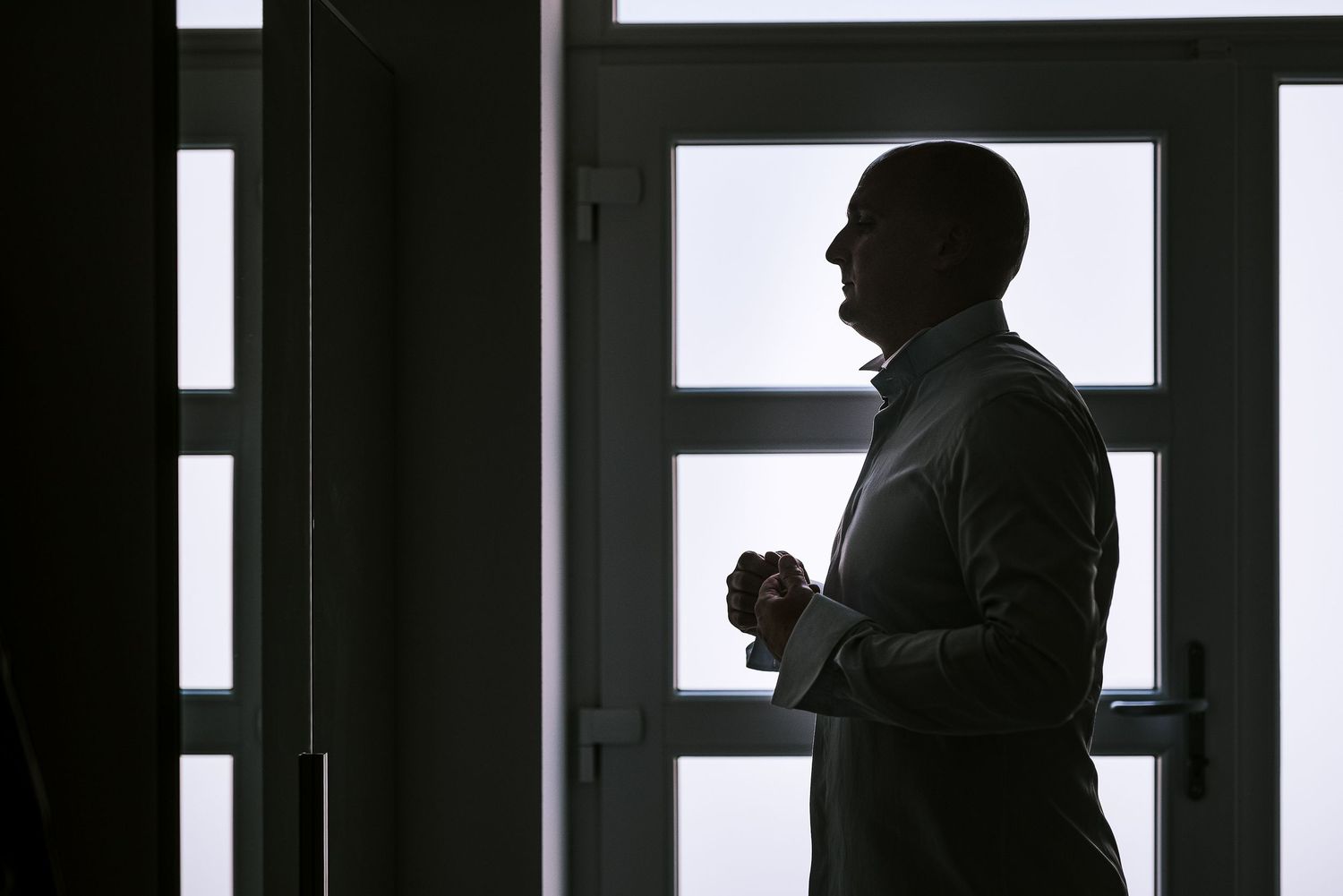 Silhouette of a man in a dress shirt standing by a window holding cufflinks while getting ready.