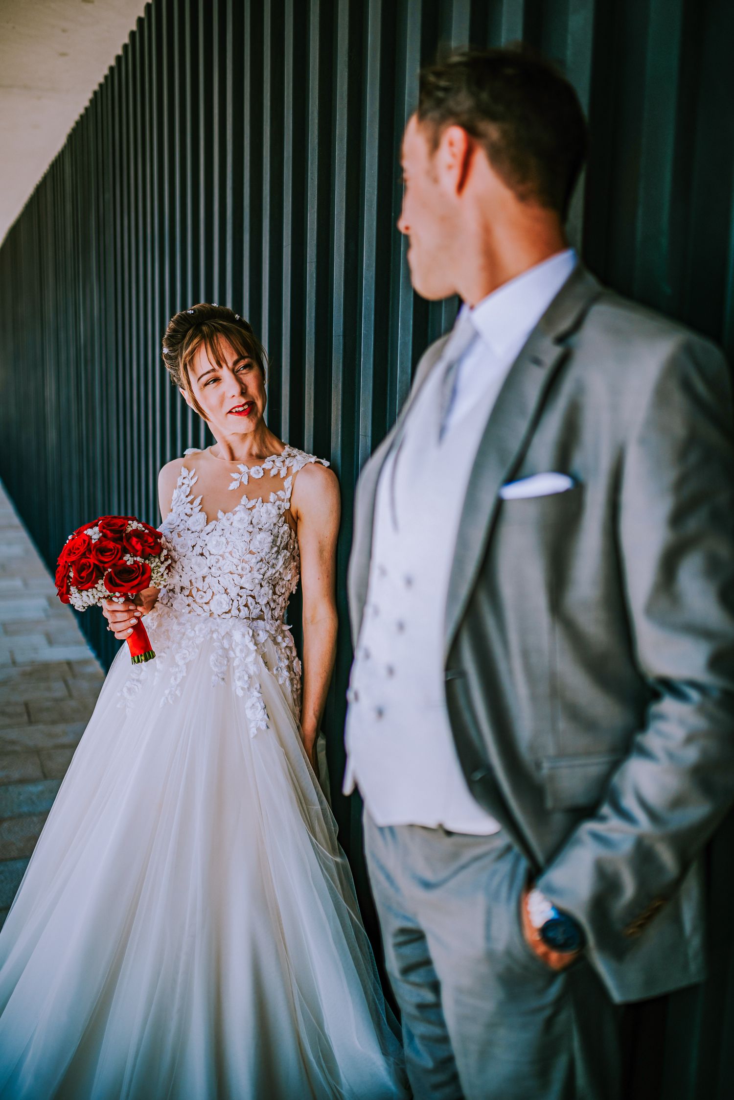 Bride in lace gown holds red bouquet gazing at groom in gray suit against striped wall.