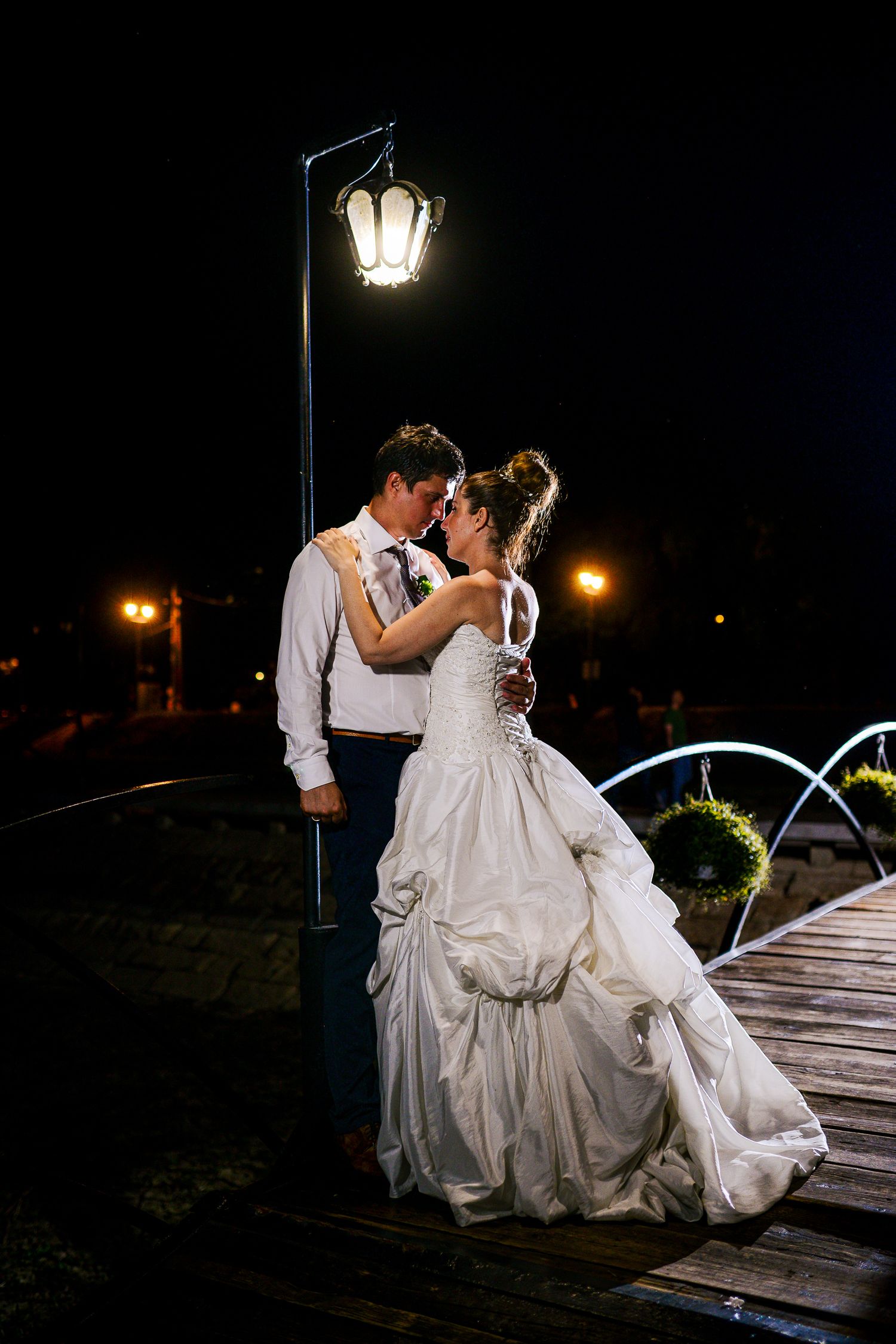 Bride and groom share romantic embrace under glowing street lamp on wooden bridge at night.