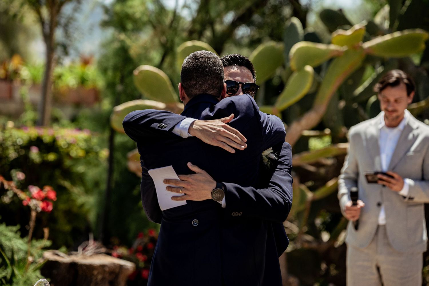 Two men in suits share an emotional hug at an outdoor wedding celebration surrounded by greenery.