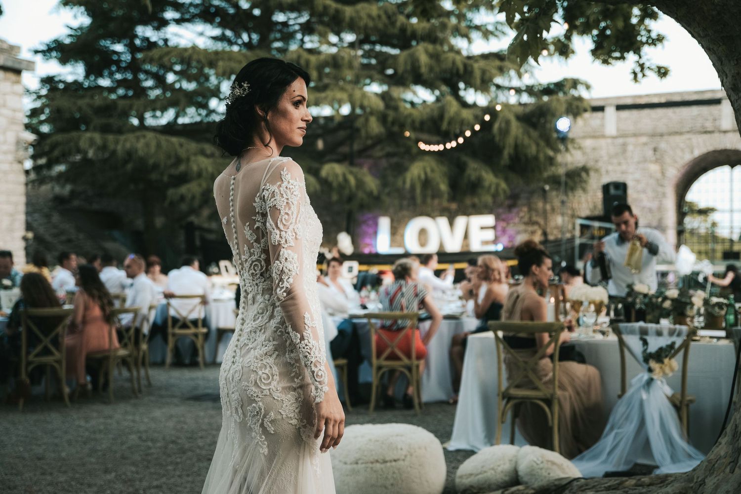 Bride in lace gown standing at outdoor wedding reception with illuminated LOVE sign in background.