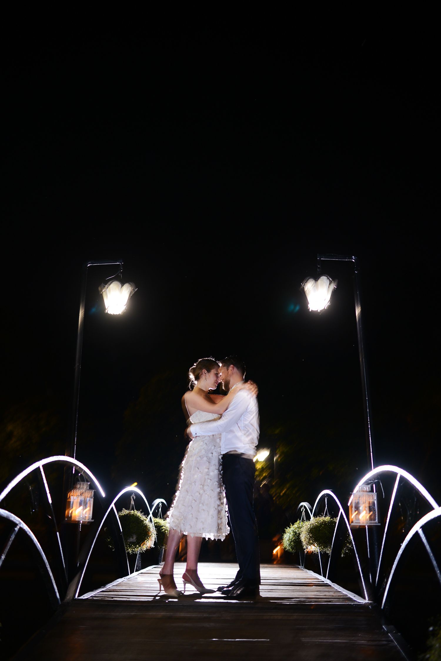 Bride and groom share a romantic kiss on a lit bridge at night during their wedding celebration.