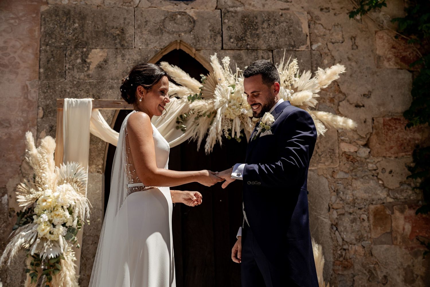 Bride and groom exchange rings before a stunning pampas grass arch at rustic outdoor wedding.