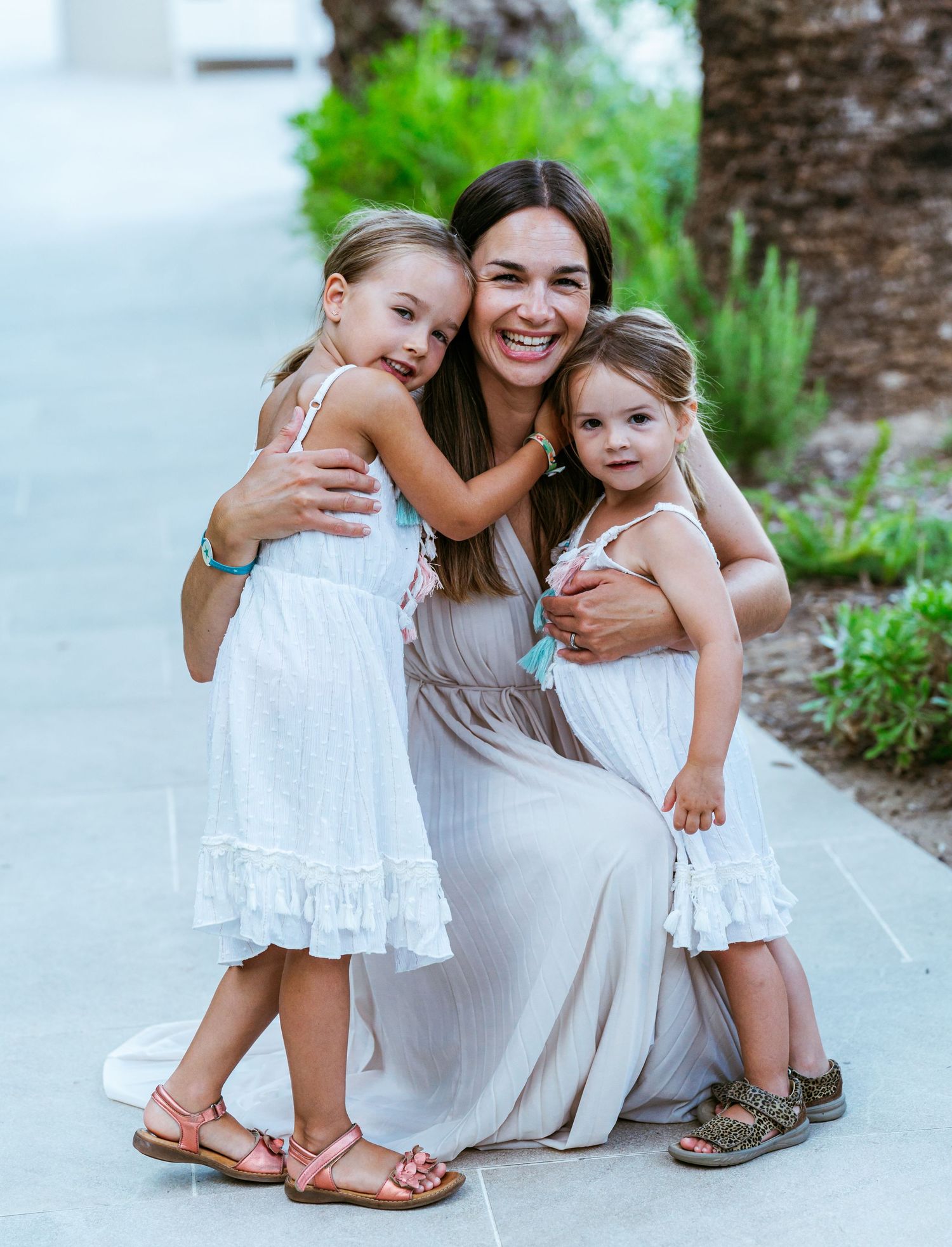 Smiling mother crouches down as her two young daughters in white dresses hug her warmly outdoors.