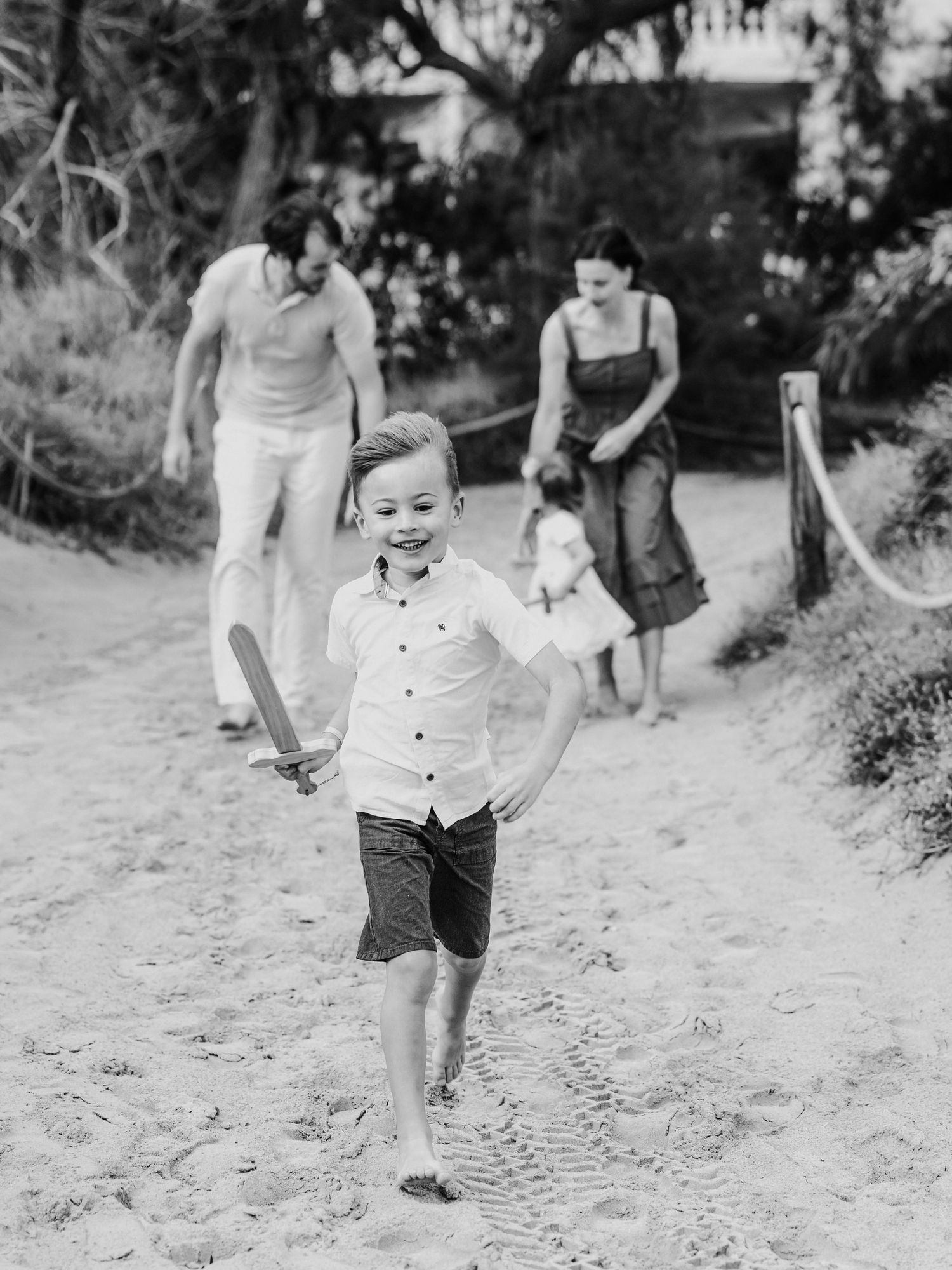 Black and white photo of smiling boy running on beach with family walking behind him.