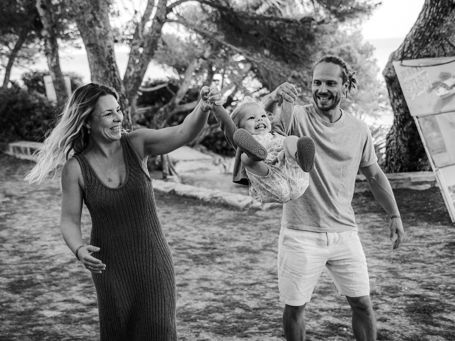 Black and white photo of happy family swinging toddler between parents outdoors among pine trees.