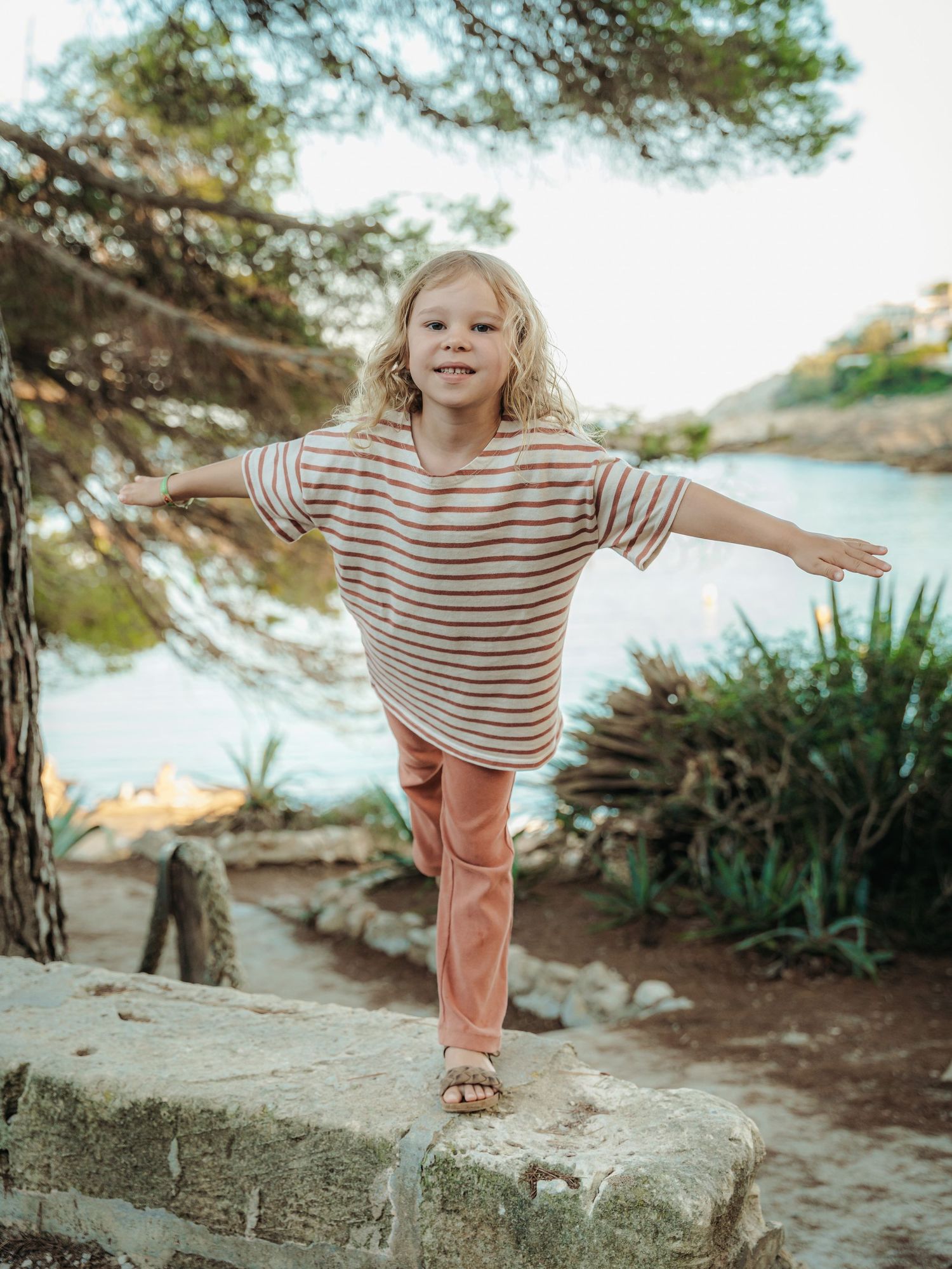 Cheerful blonde girl in striped top balancing on a stone wall with arms out near a scenic cove.