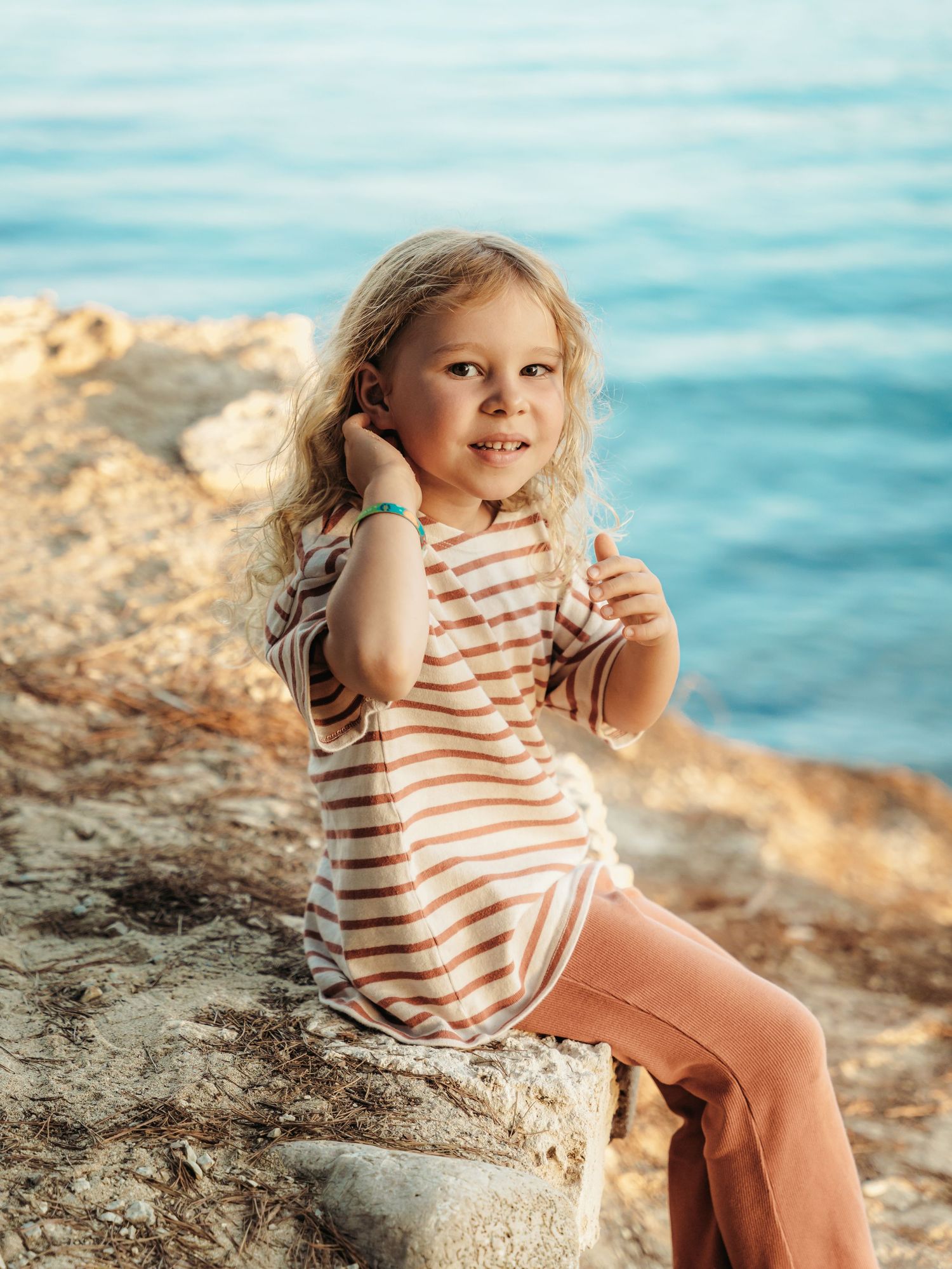 Young blonde girl in striped shirt sitting on rocks by turquoise water, smiling.