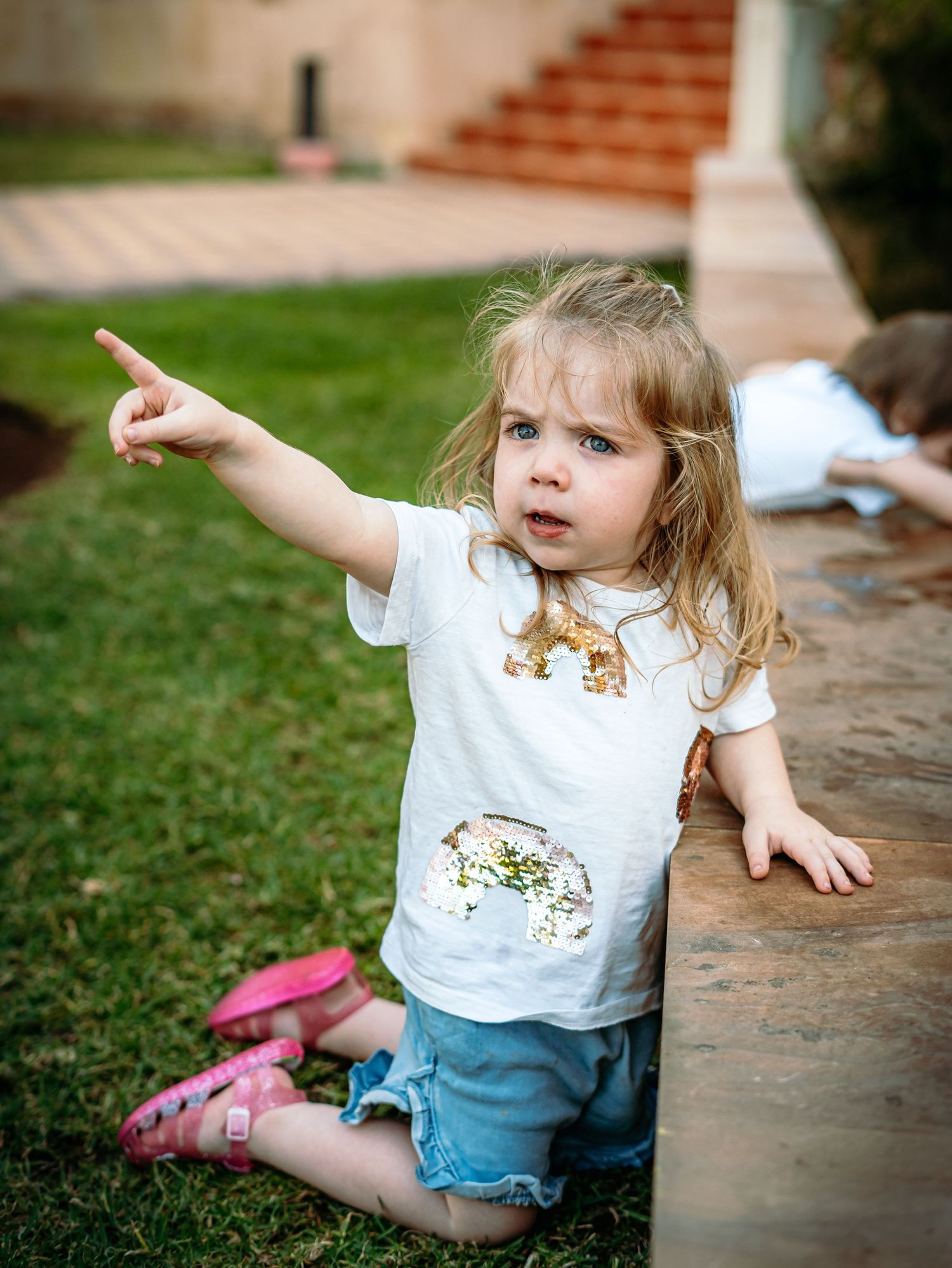 Curious blonde toddler girl in rainbow sequin shirt pointing finger in garden.