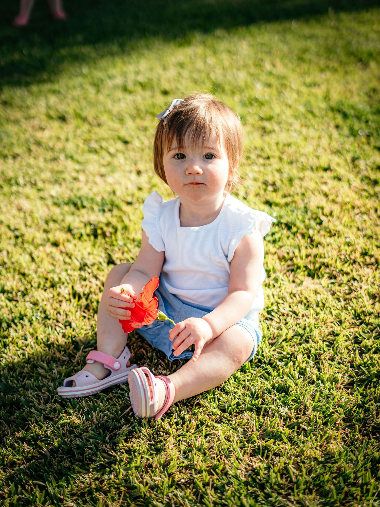 Cute baby girl sitting on green grass outdoors holding a red toy wearing white top and blue skirt.
