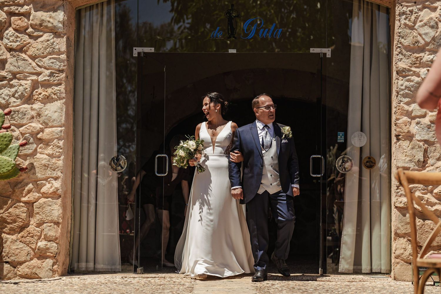 Bride in elegant white gown with bouquet walks arm-in-arm with escort through stone venue entrance.