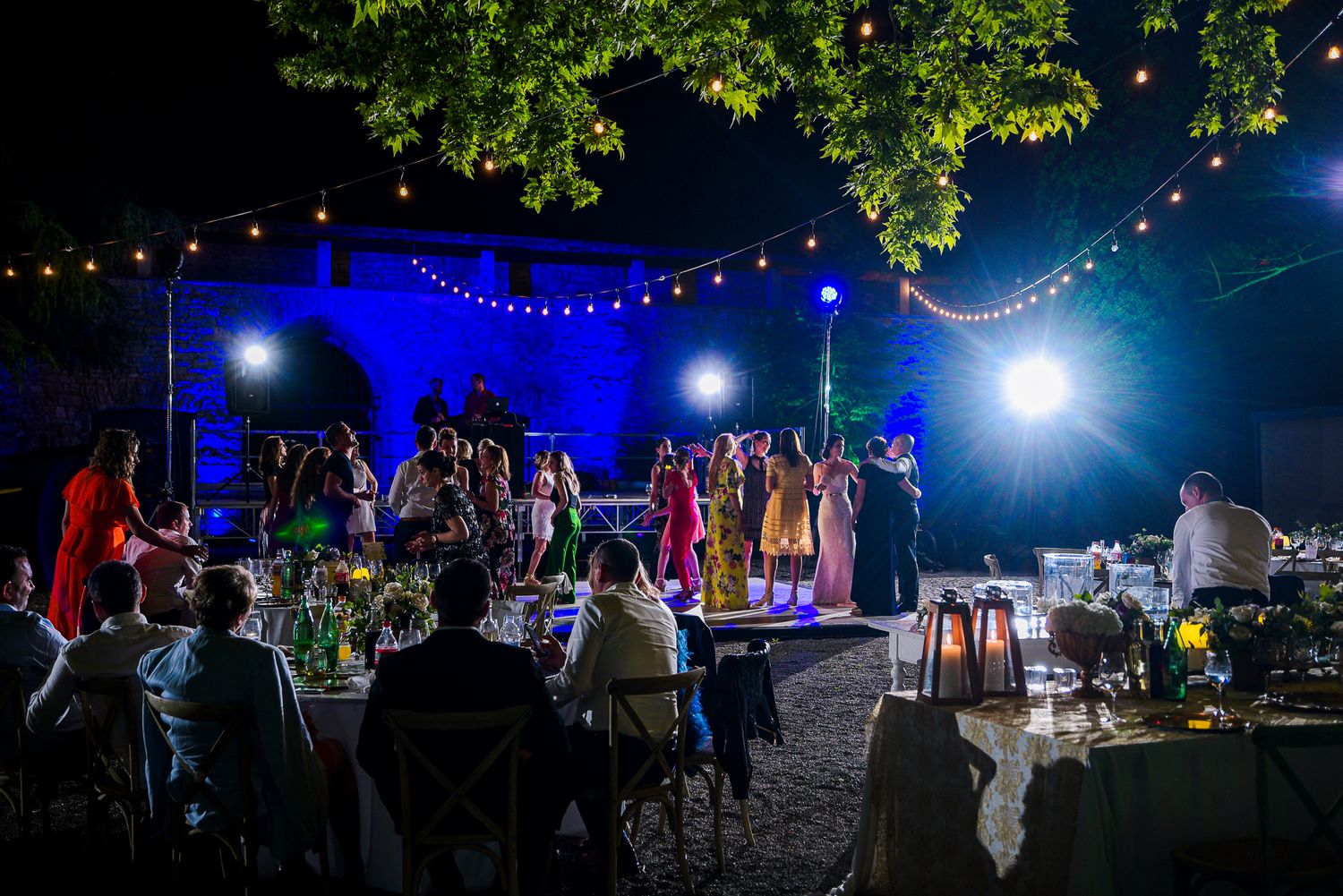 Outdoor nighttime wedding reception with string lights, guests dancing, and blue stage lighting.