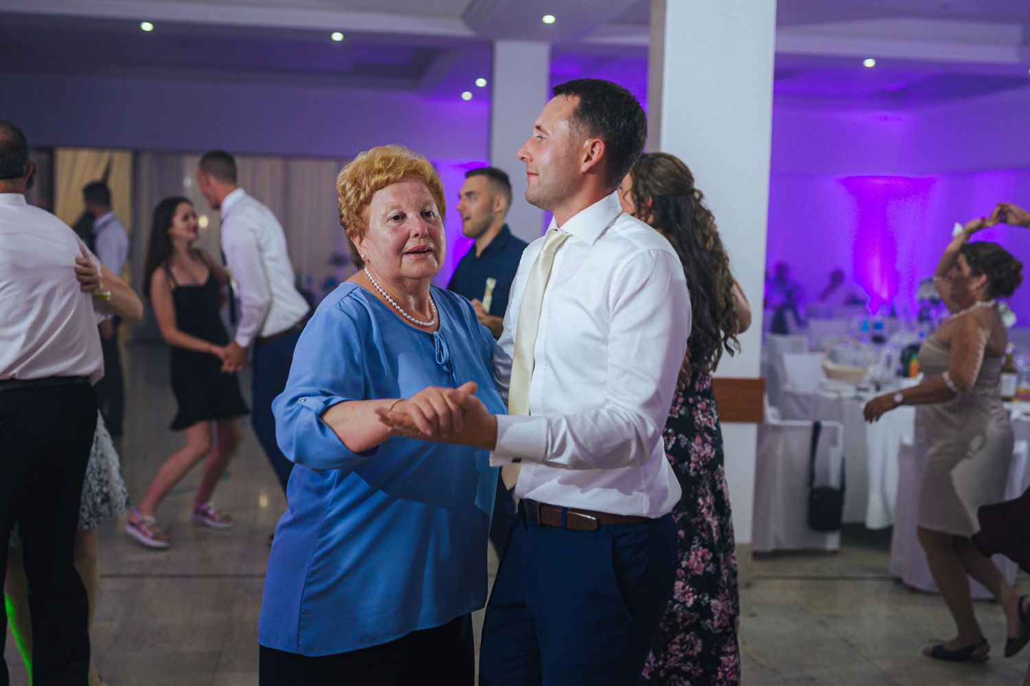 Elderly woman in blue dress dances with man in white shirt at wedding reception.