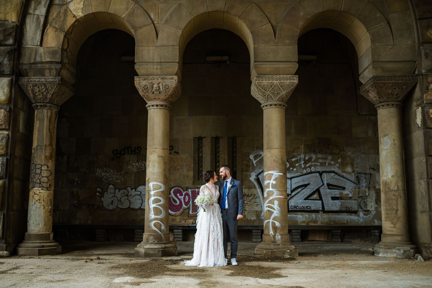 Bride and groom standing together in a grand abandoned building with arched columns and graffiti walls.