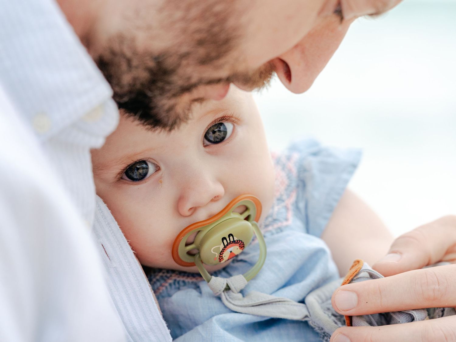 Father tenderly holds baby with pacifier in blue outfit, close-up portrait.