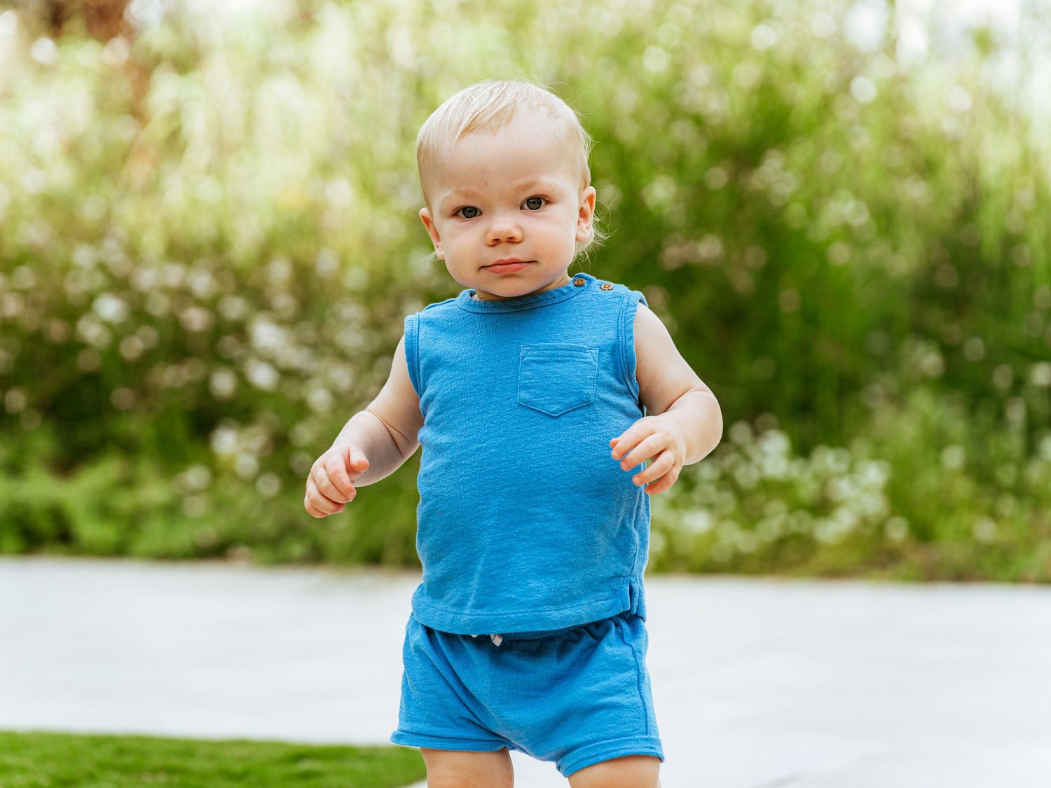Adorable toddler in a blue outfit walking outdoors in a lush green garden setting.