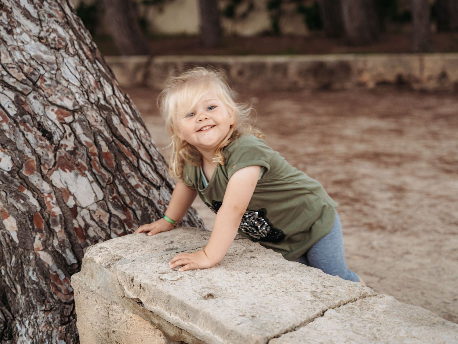 Adorable blonde toddler girl in green shirt climbing on stone wall next to large tree trunk.