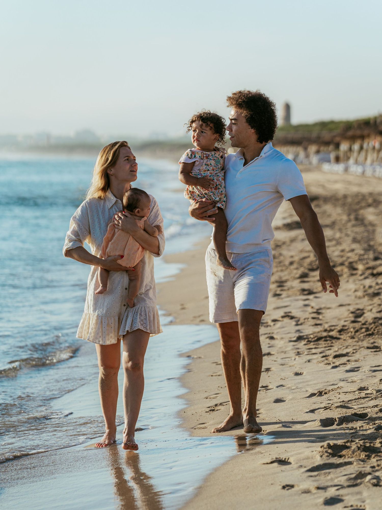 A young family walks barefoot on a sunny beach, parents each holding a small child near the ocean waves.