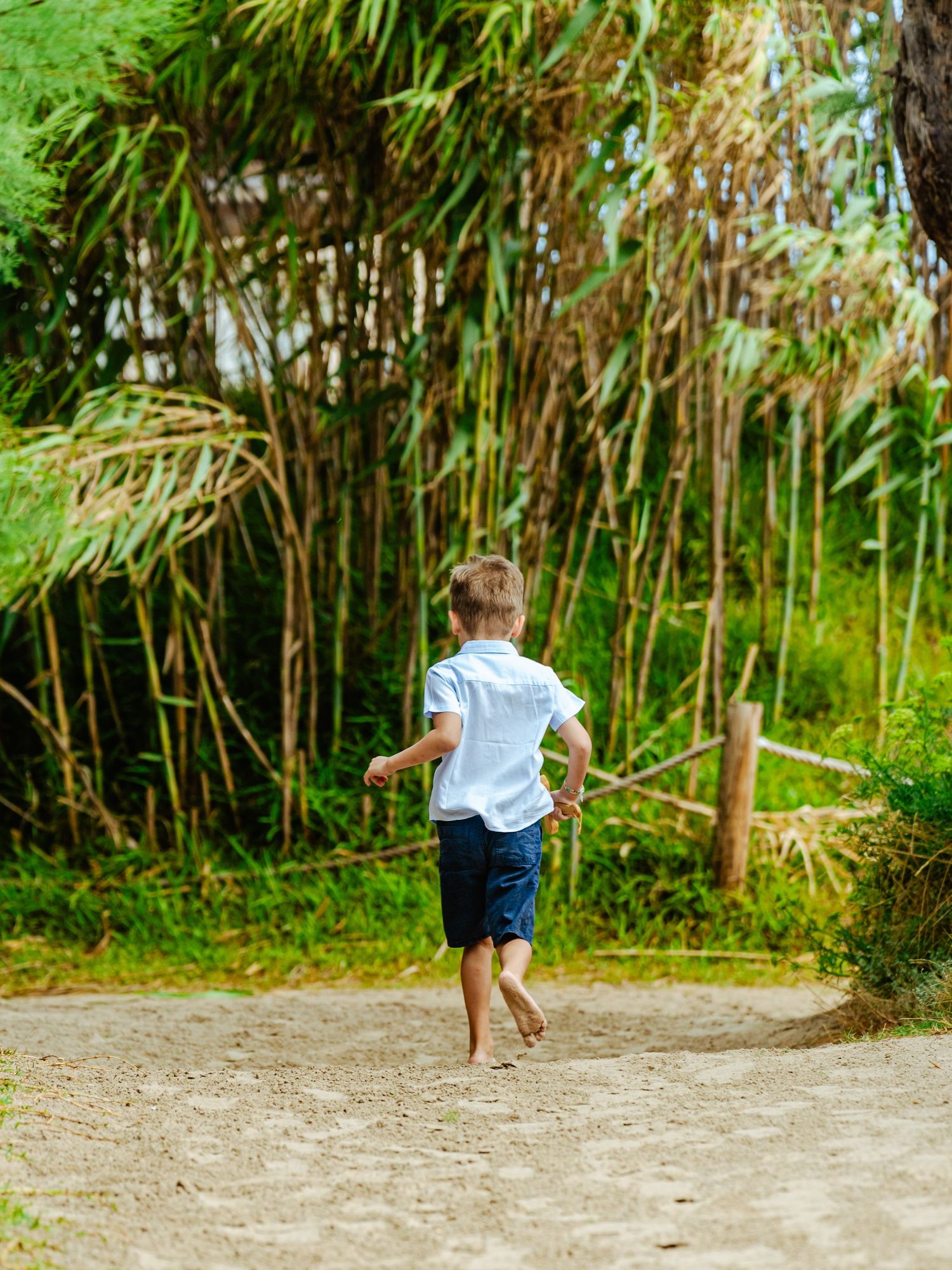 Barefoot young boy in white shirt and blue shorts running along a sandy path surrounded by tropical greenery.