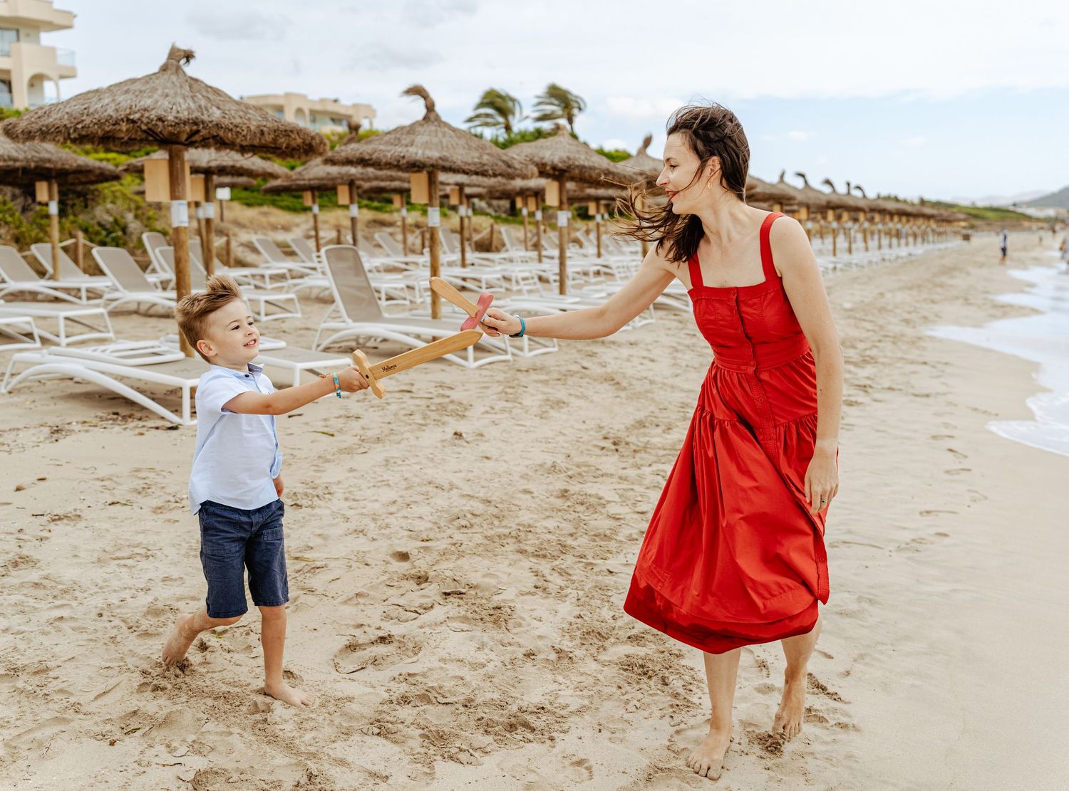 Mother in red dress playing with young son on sunny sandy beach.