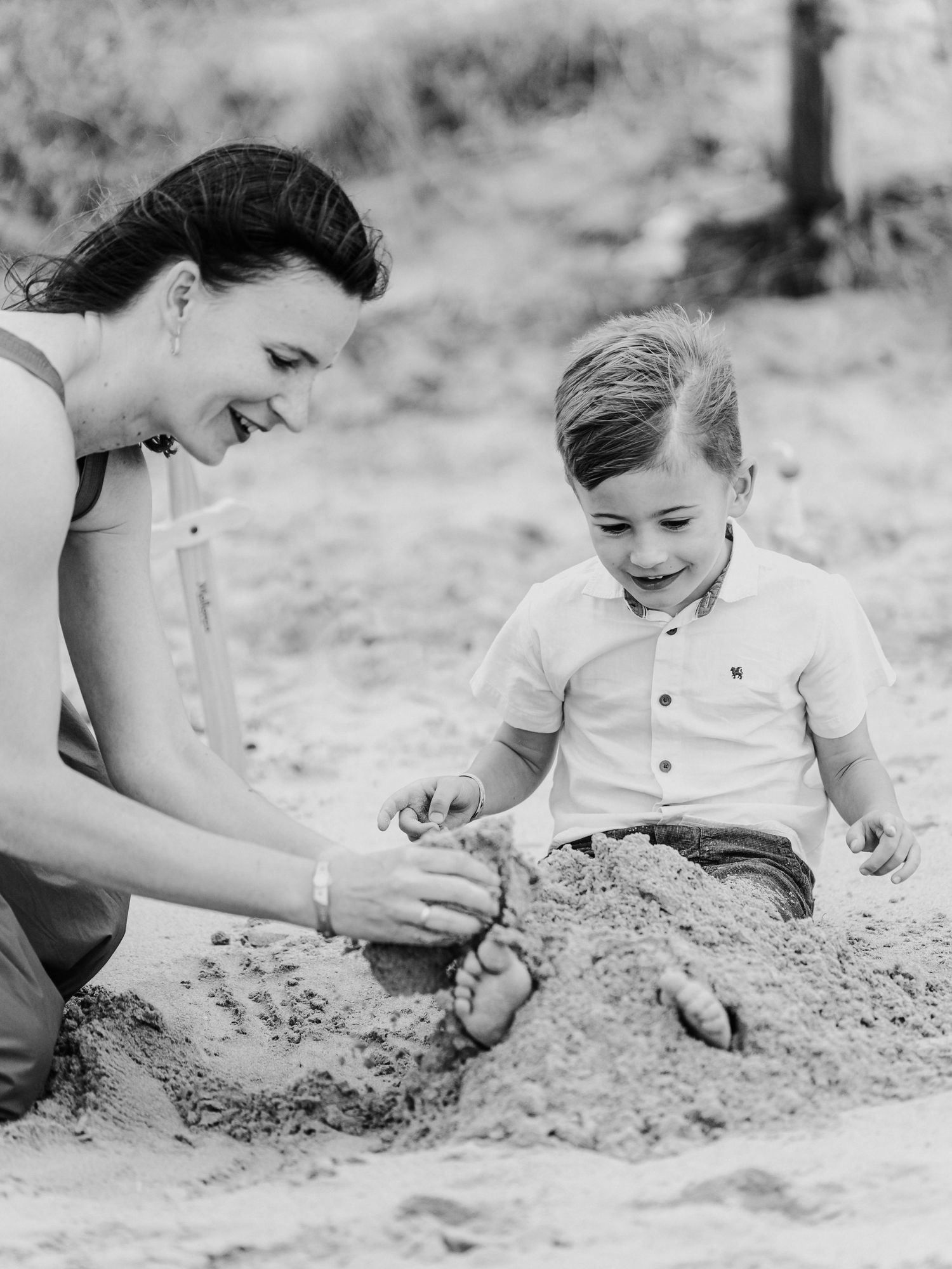 Black and white photo of a smiling mother and young boy playing in sand at the beach.