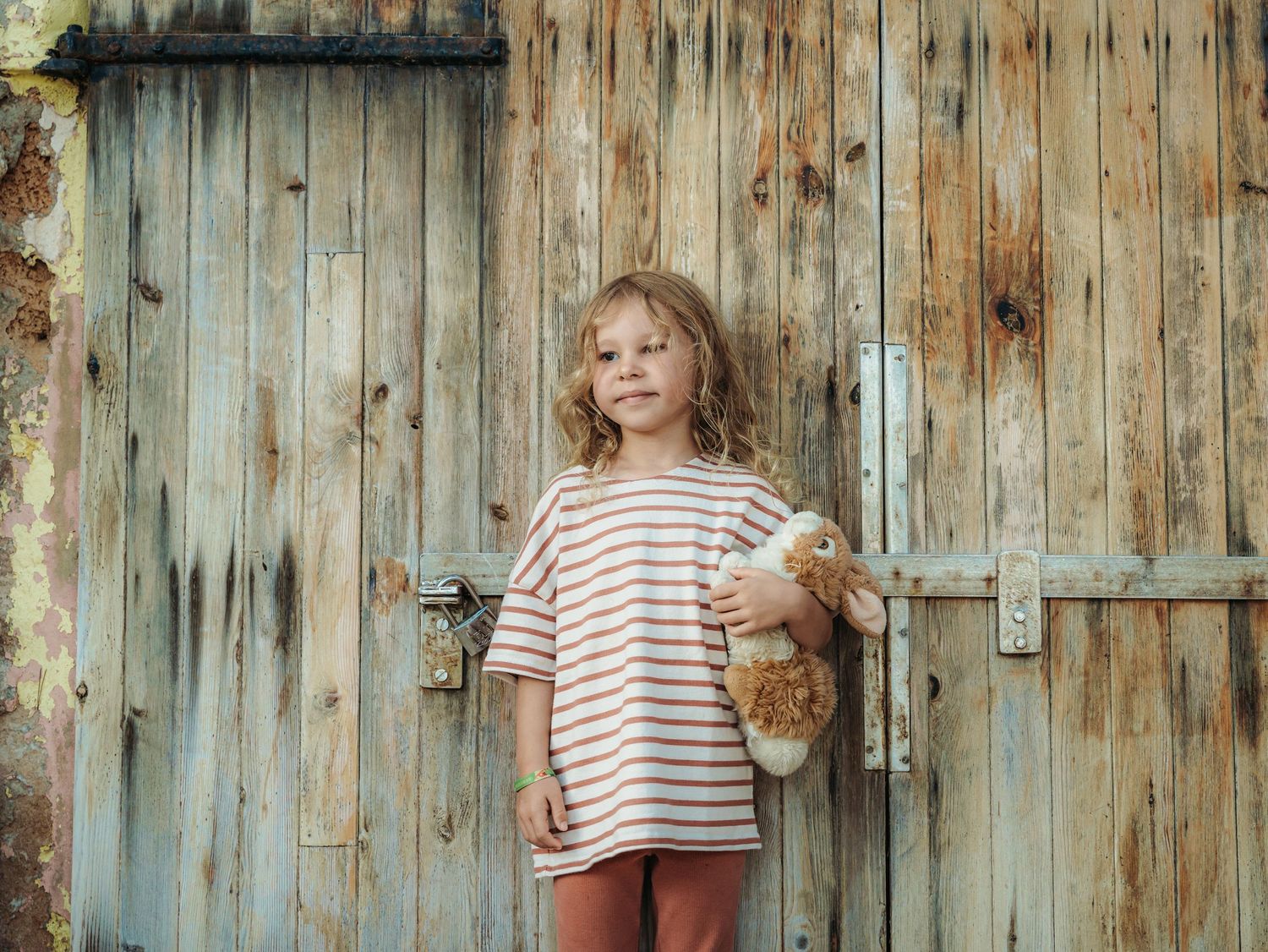 Young girl in striped oversized tee holding teddy bear standing before rustic weathered wooden door.