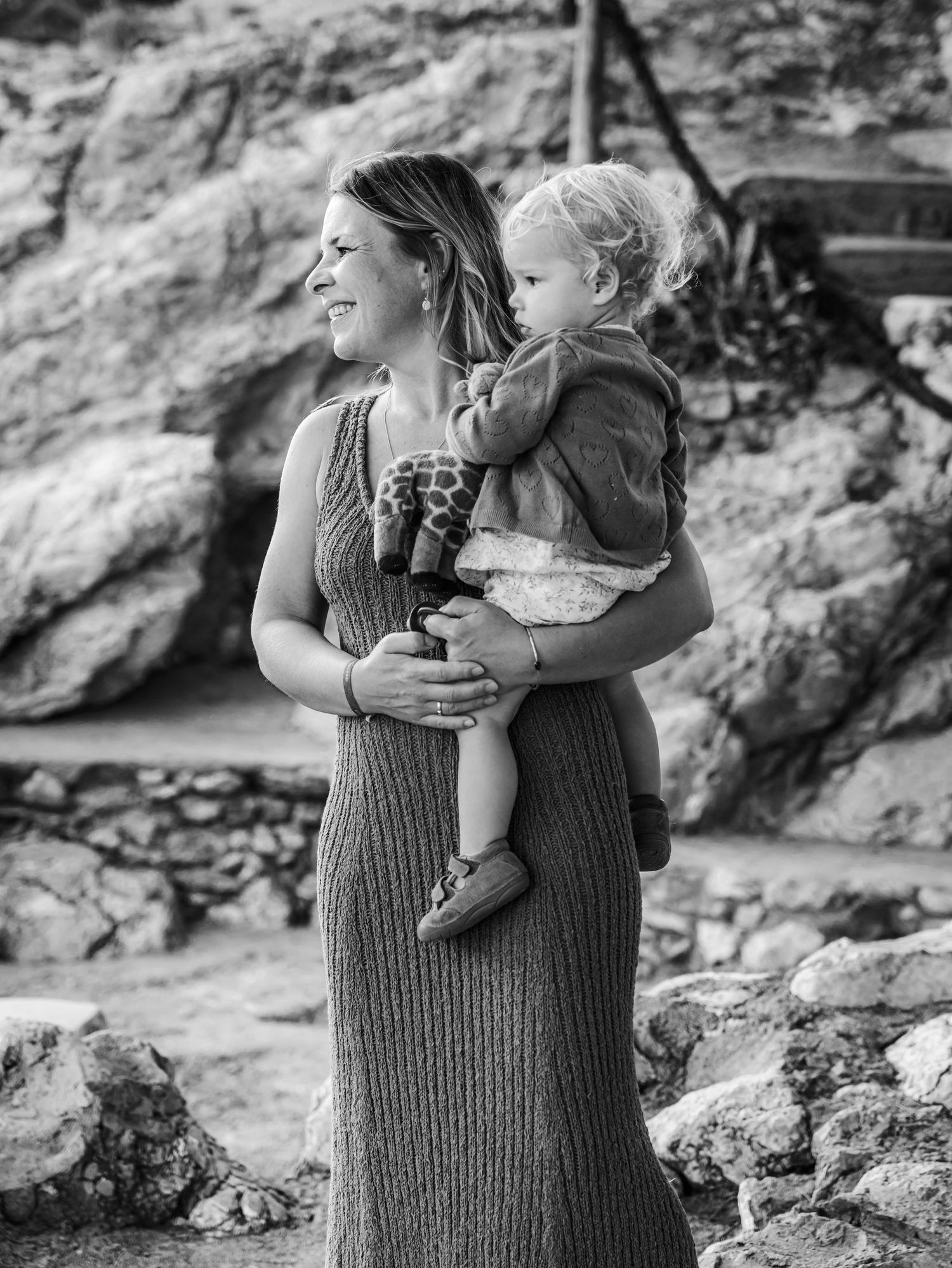 Black and white photo of a smiling mother holding her toddler against a rocky outdoor backdrop.