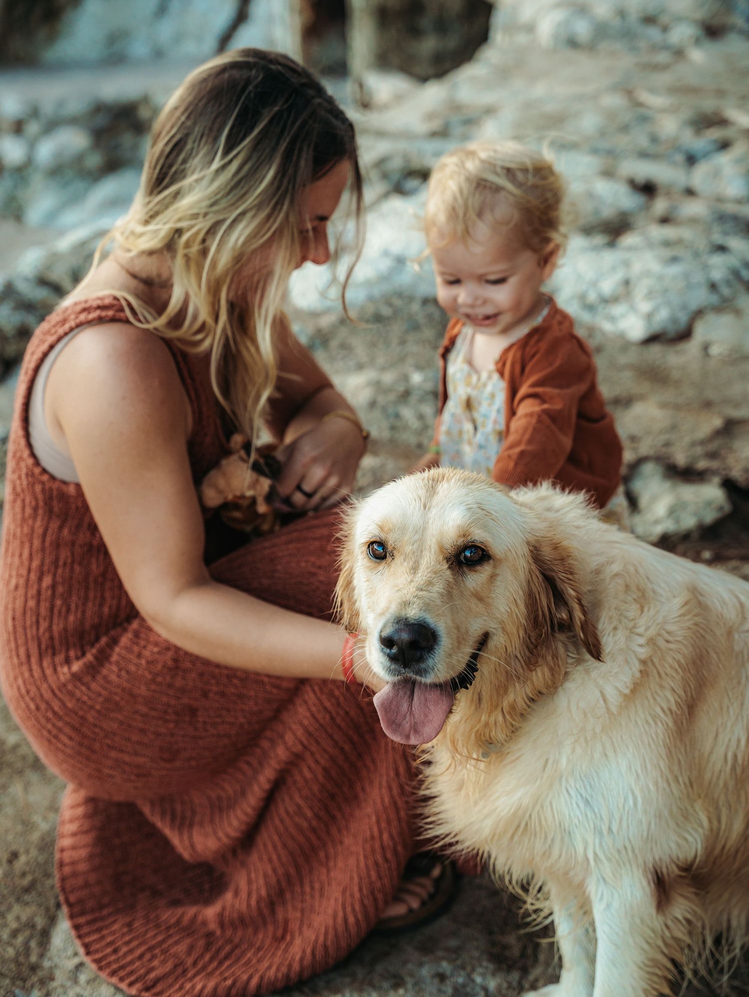 A woman in rust dress pets a golden retriever while a toddler in brown outfit watches near rocky shore.