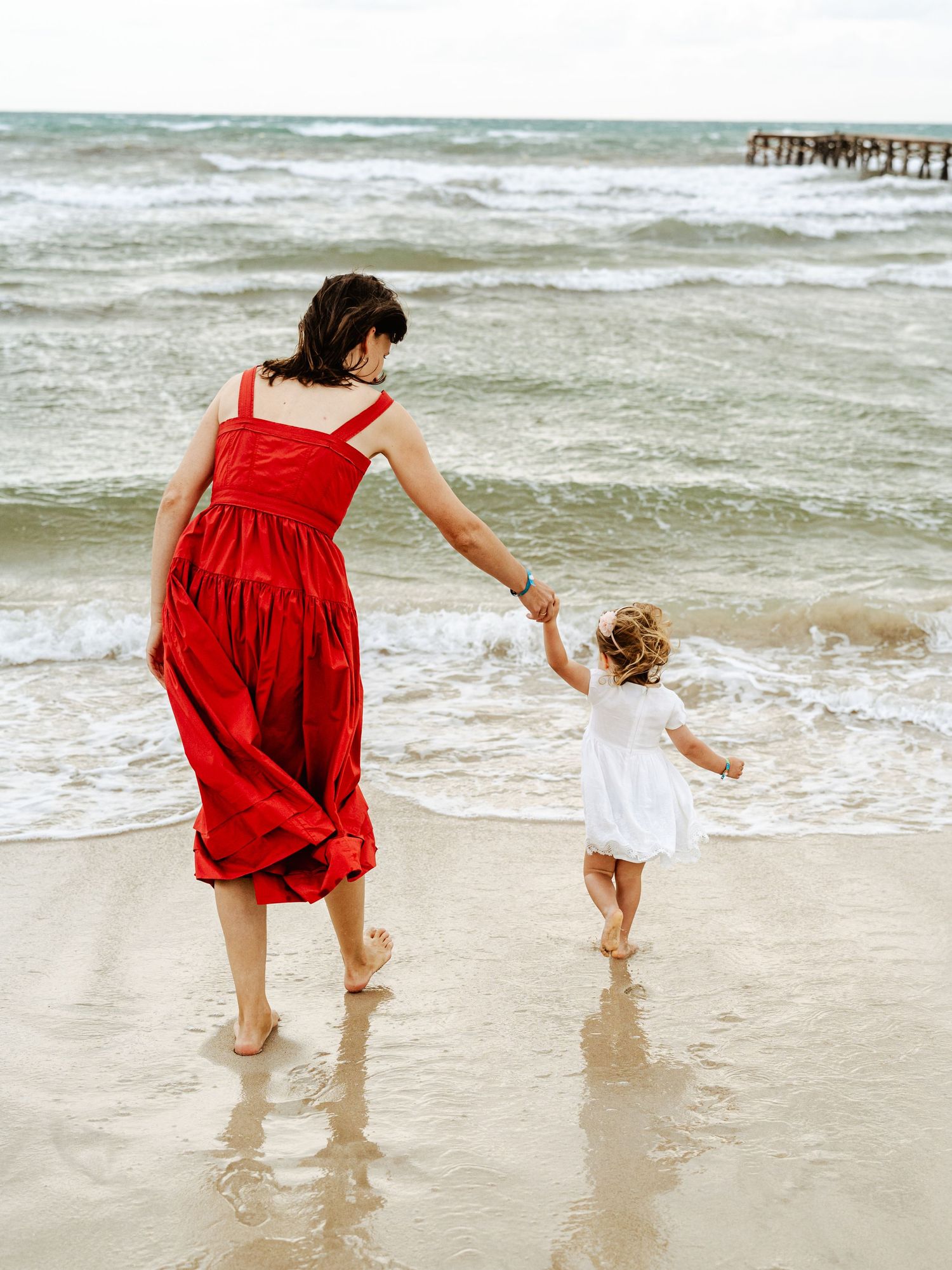 Mother in red dress leads young child in white dress along a sandy ocean shoreline.