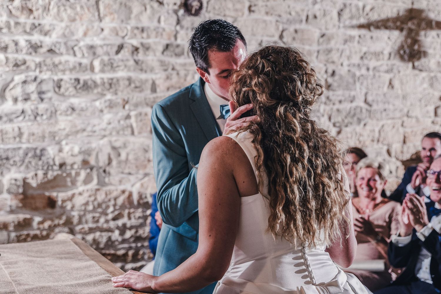 Bride and groom sharing their first kiss during wedding ceremony against a rustic stone wall.