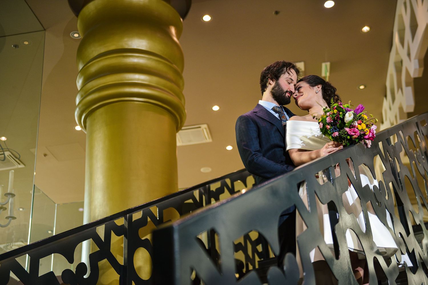 Bride and groom share a romantic moment on a staircase beside a gold column at their wedding venue.