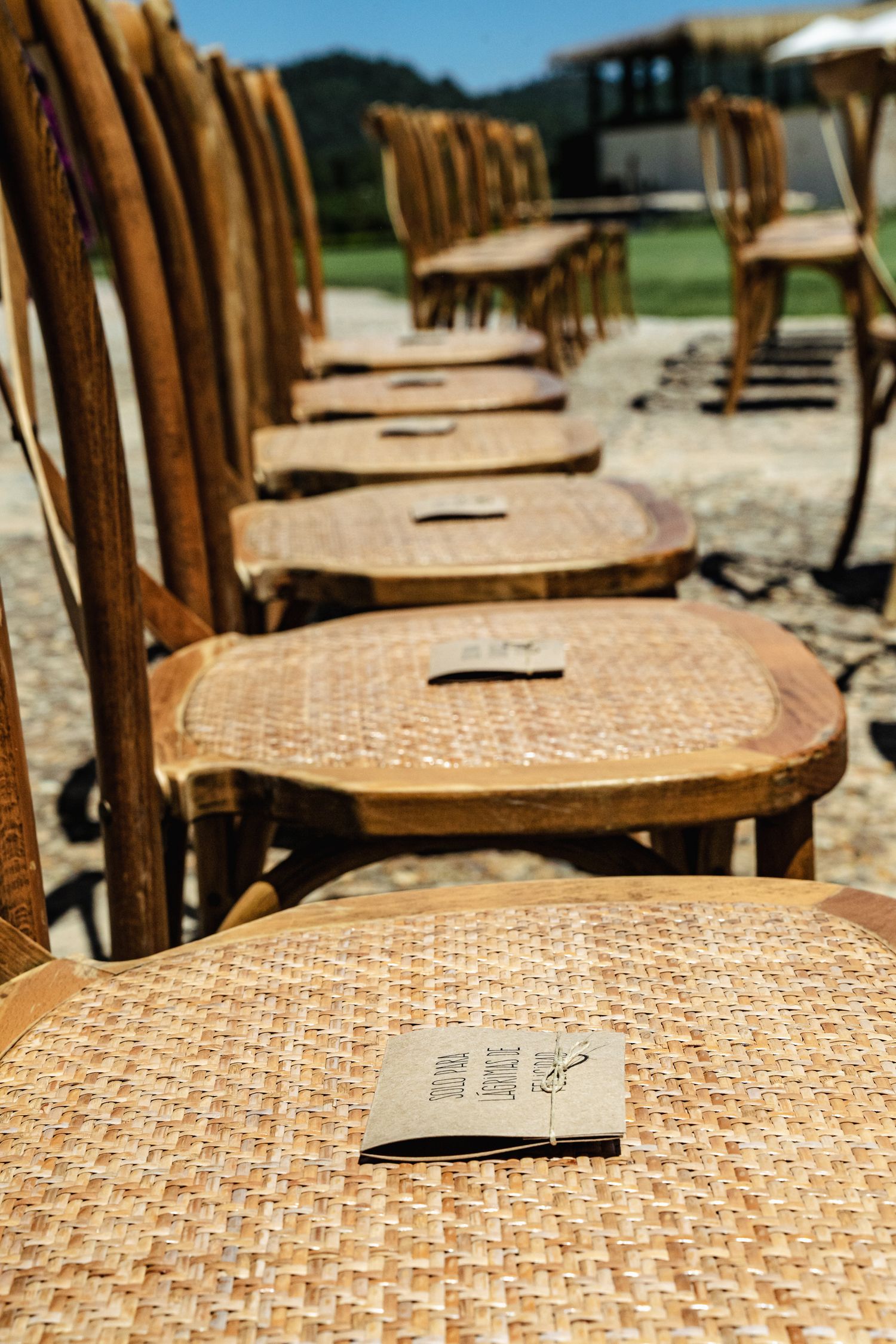 Rows of rustic wooden rattan chairs set up outdoors for a wedding ceremony with programs placed on seats.