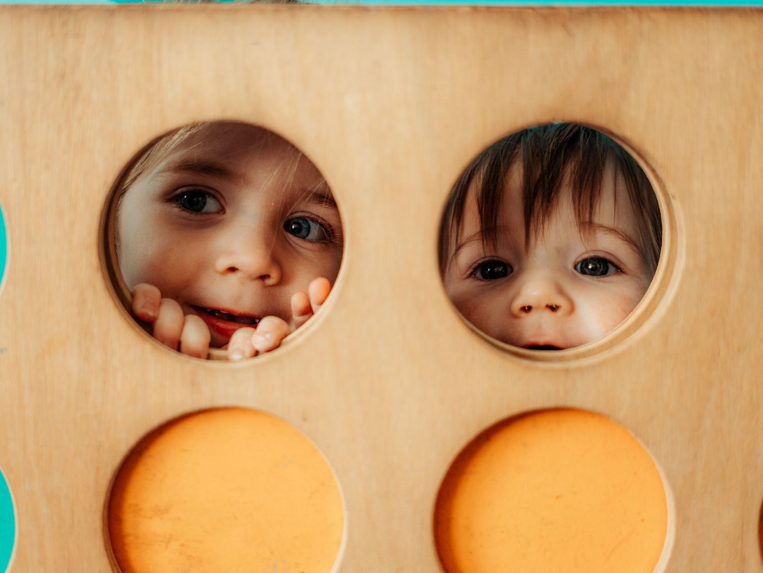 Two sisters peeking playfully through circular openings in a wooden play structure during a candid family photography session.