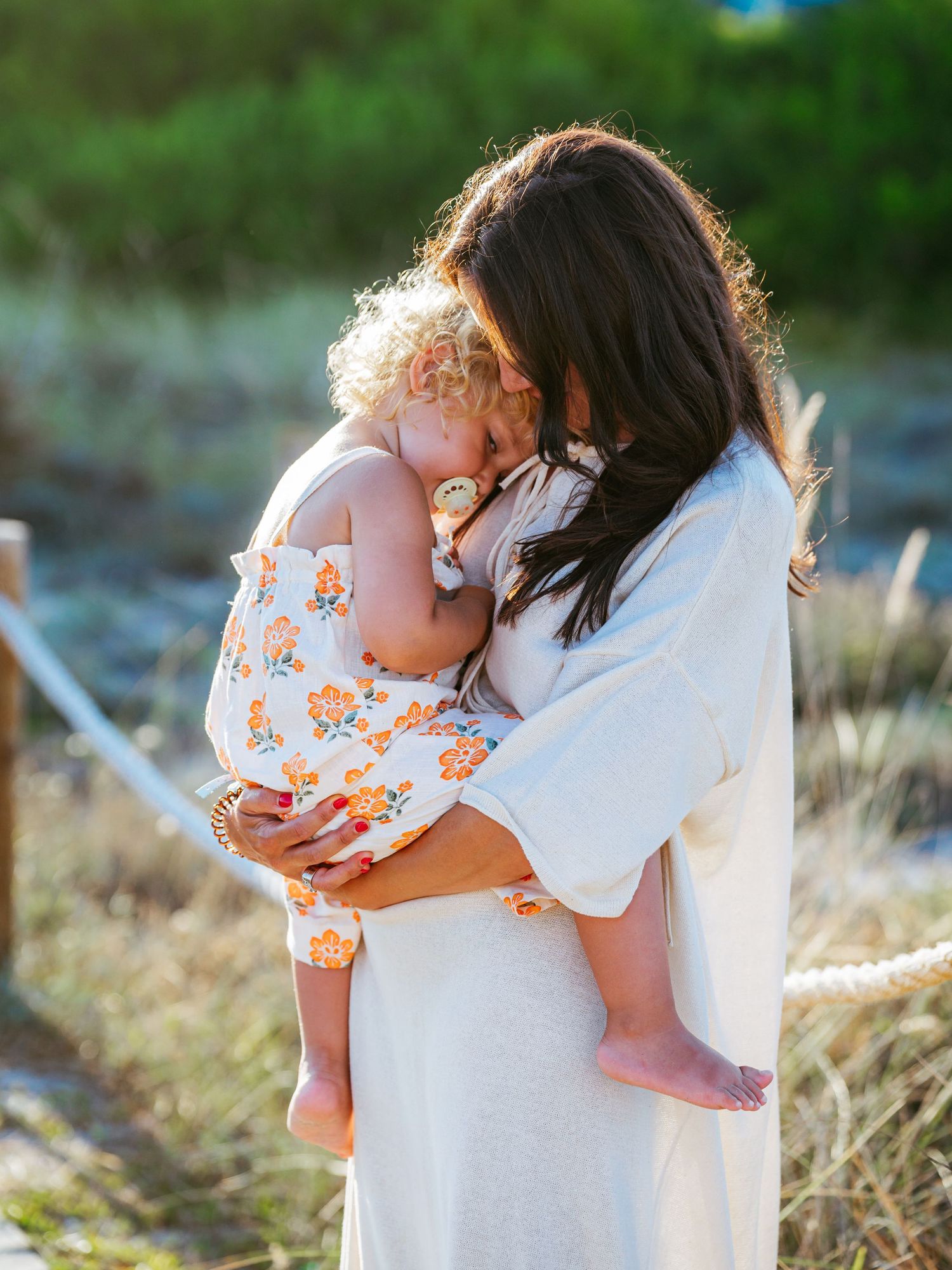 Loving dark-haired mother kissing her curly-haired toddler in a floral romper at a sunny beach.