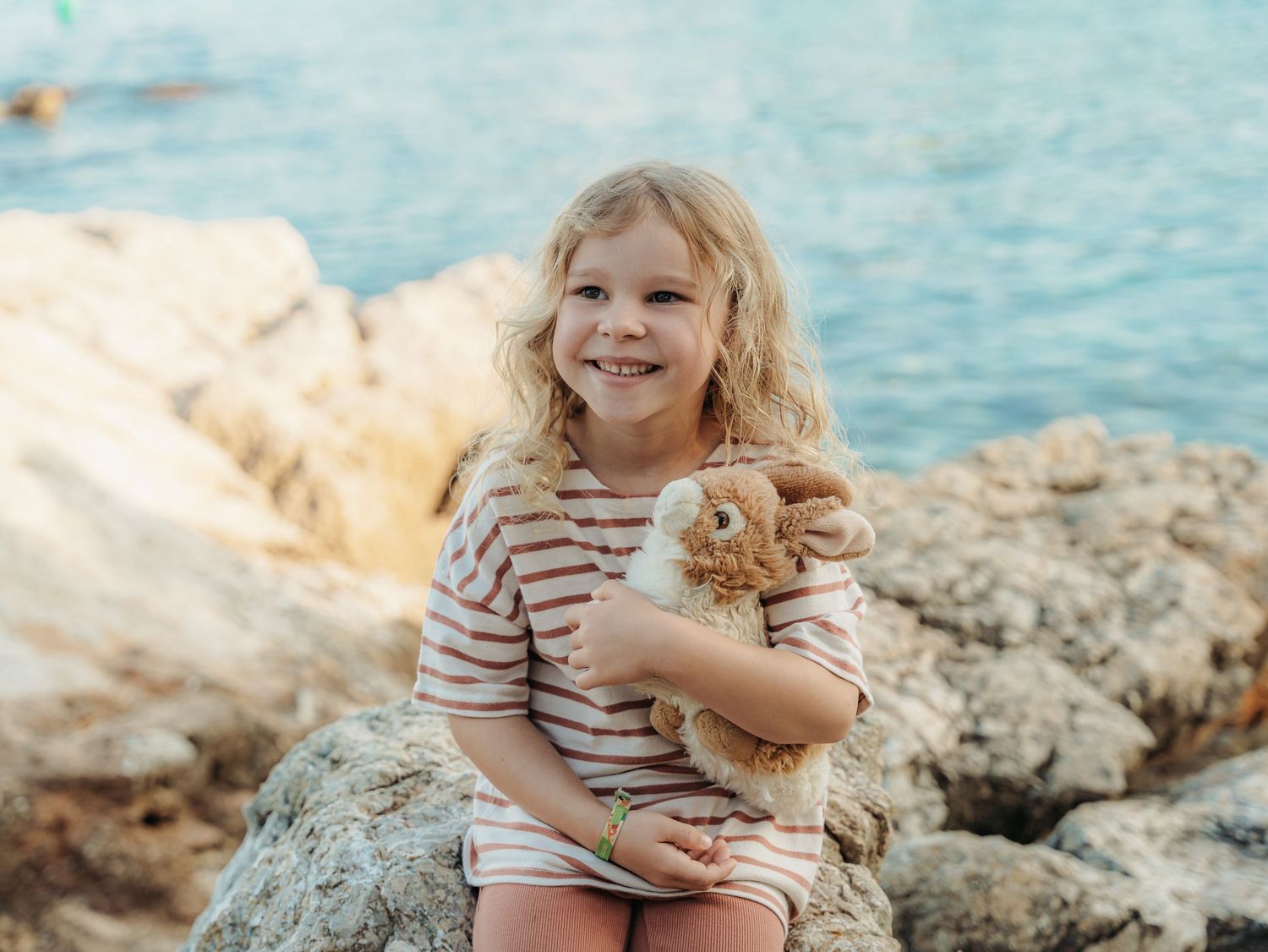 Smiling young girl with blonde curls hugging a plush bunny while sitting on seaside rocks.