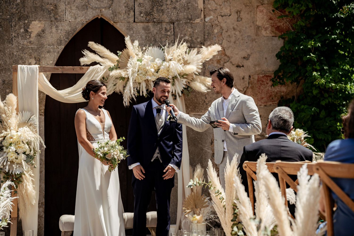 Bride and groom exchange vows at outdoor ceremony with elegant pampas grass arch at stone chapel.