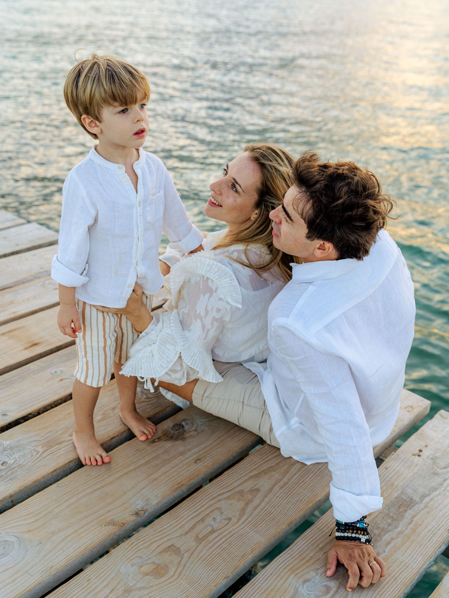 Family of three sharing a tender moment on a sunlit wooden pier overlooking calm blue ocean waters.