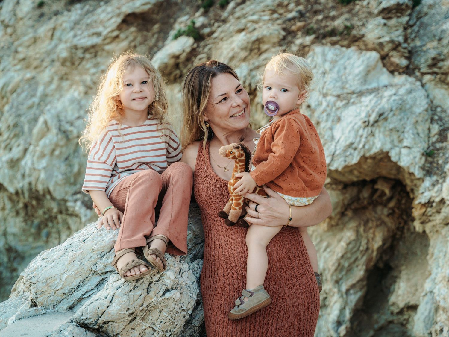 A smiling blonde woman in rust dress holds two young children against a rocky background outdoors.
