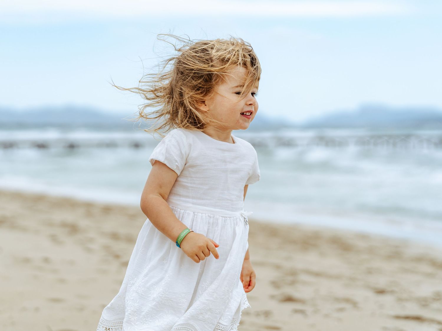 A joyful toddler girl in a white dress runs along a sandy beach with ocean waves in the background.