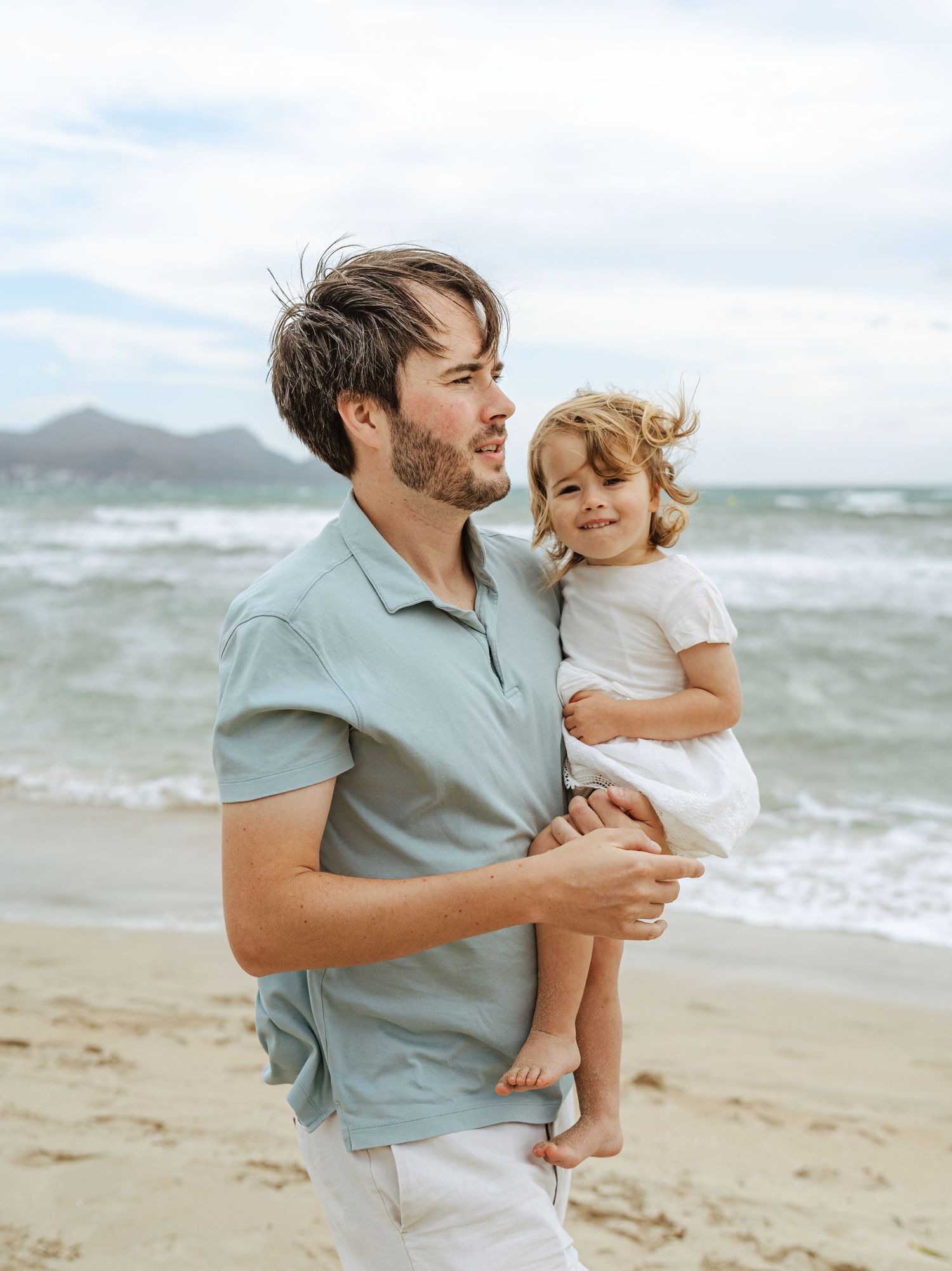 Father holding toddler girl in white dress on sandy beach with ocean waves in background.