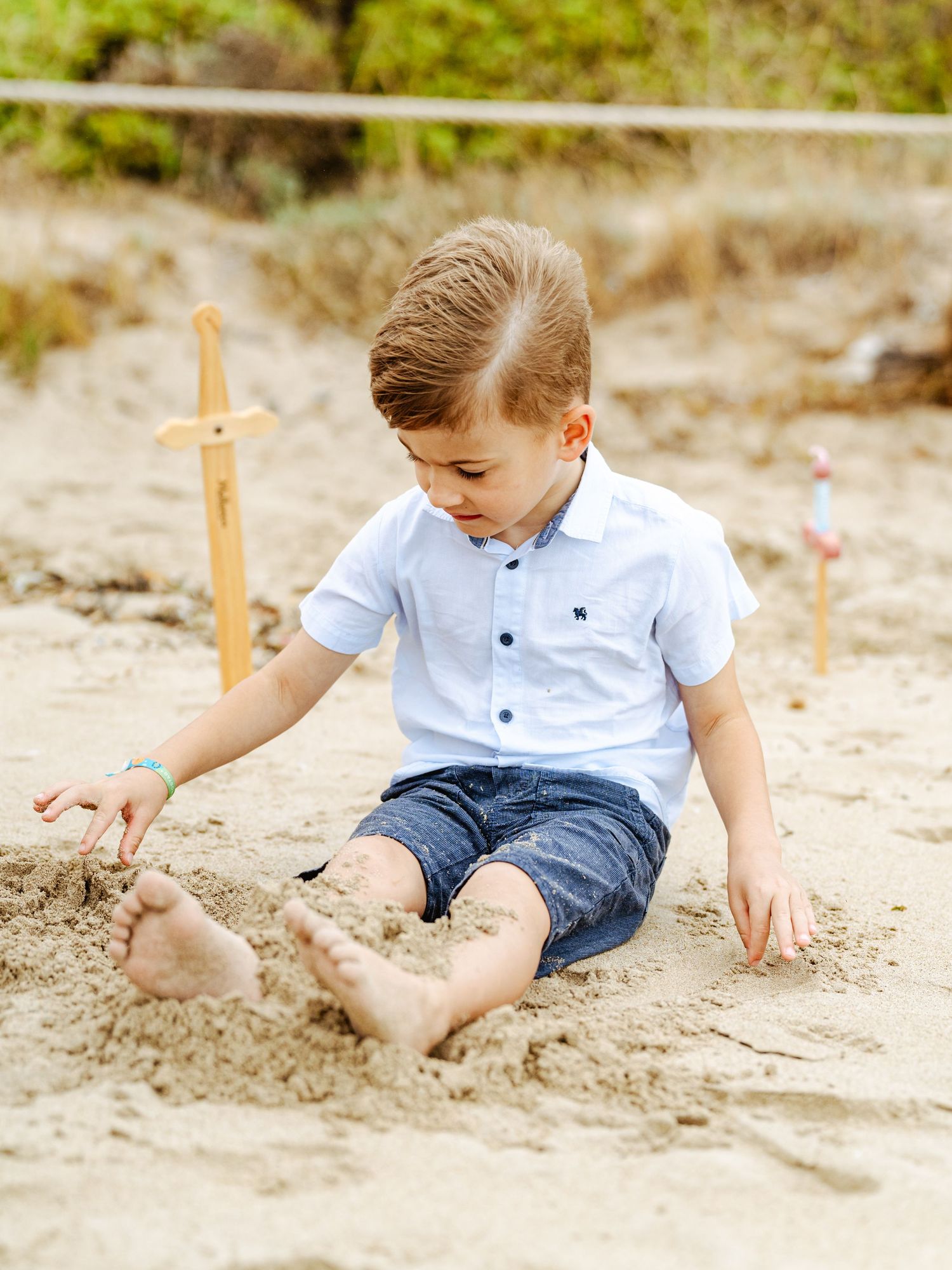 Young boy in white shirt and denim shorts playing barefoot in sand at beach.