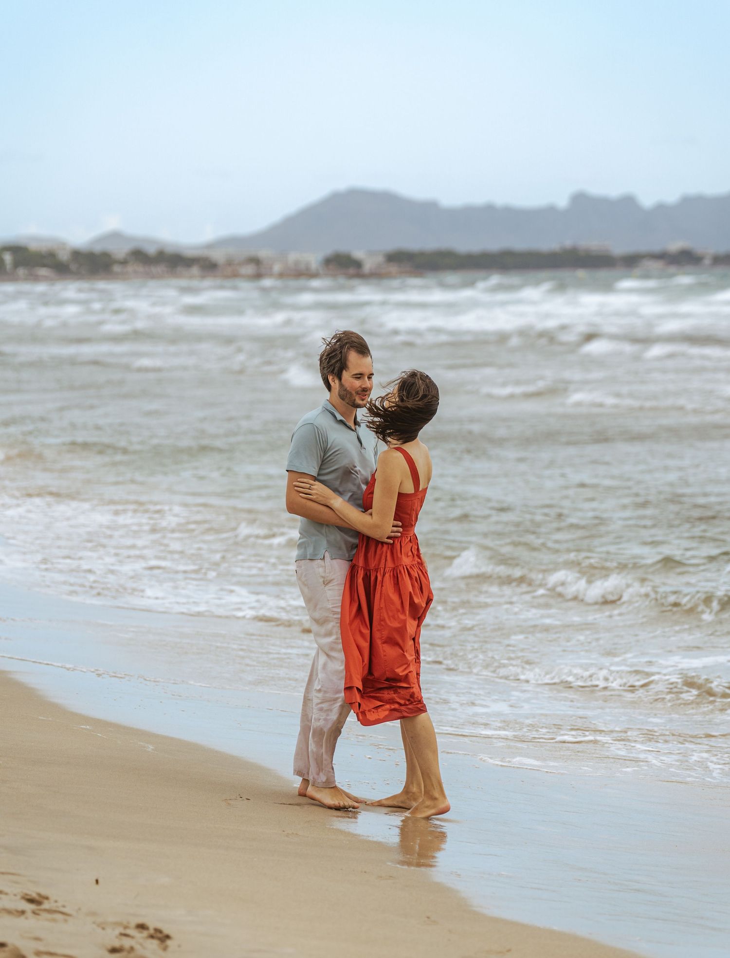 Romantic couple embracing barefoot on a sandy beach, woman in red dress, with ocean waves behind.
