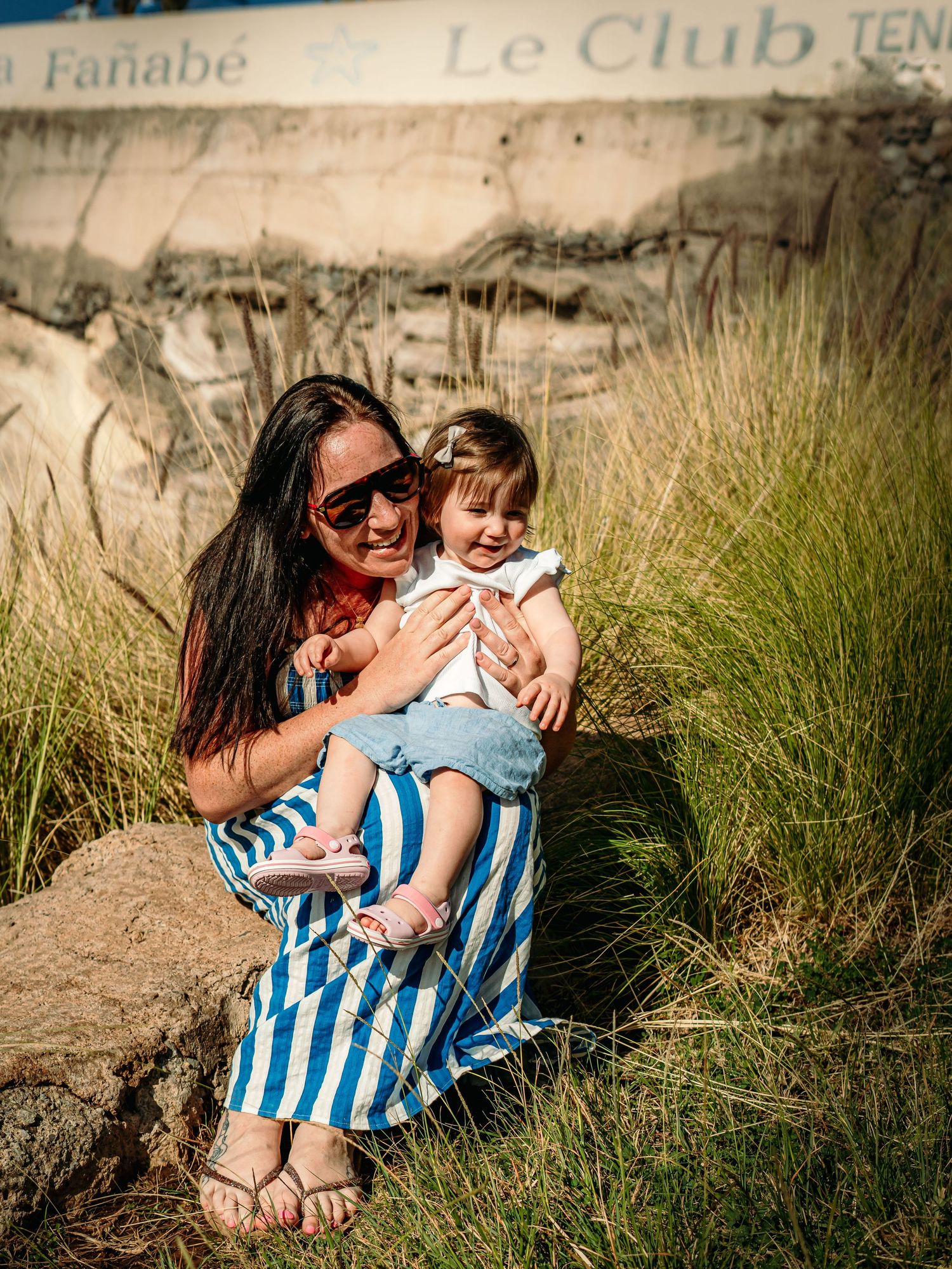 A woman in a striped dress sits on rocks holding a smiling baby near Le Club Fanabe sign.