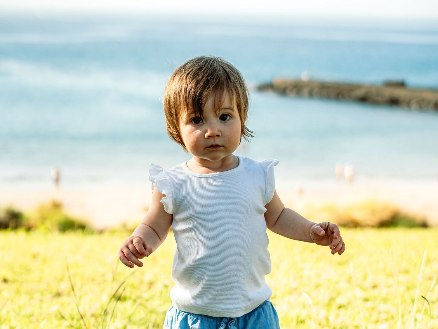 Toddler in white top and blue shorts standing in green grass with beach in background.