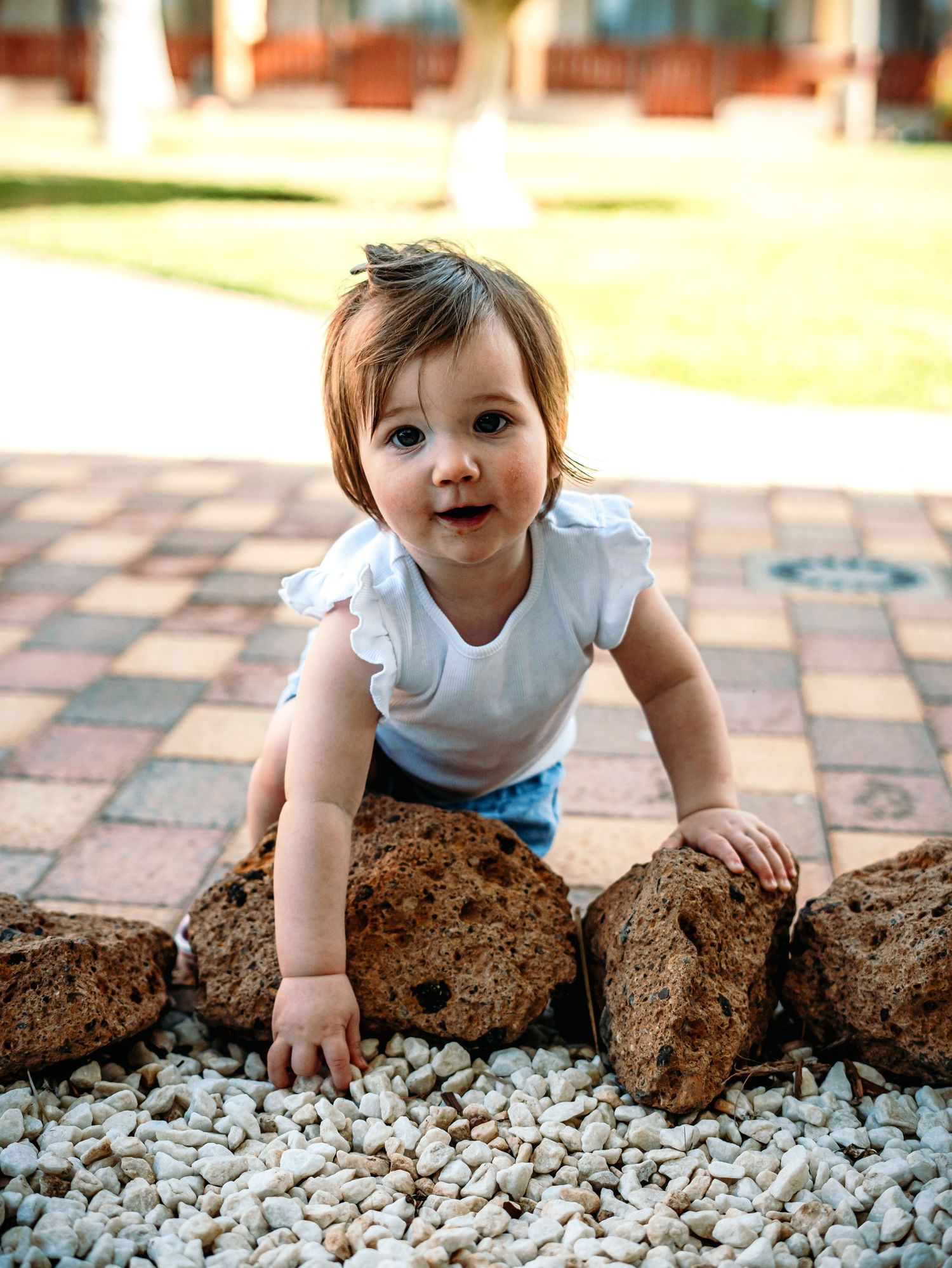 Cute baby girl in white top crawling over decorative brown rocks on white pebbles outdoors.