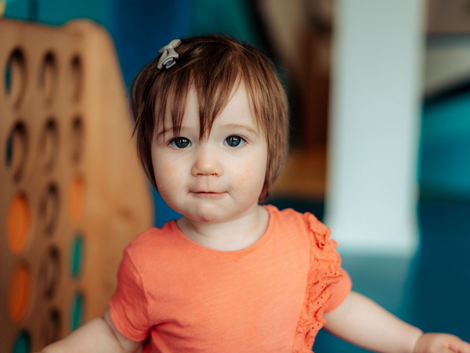 Cute toddler girl with brown hair wearing orange shirt standing near colorful play equipment indoors.
