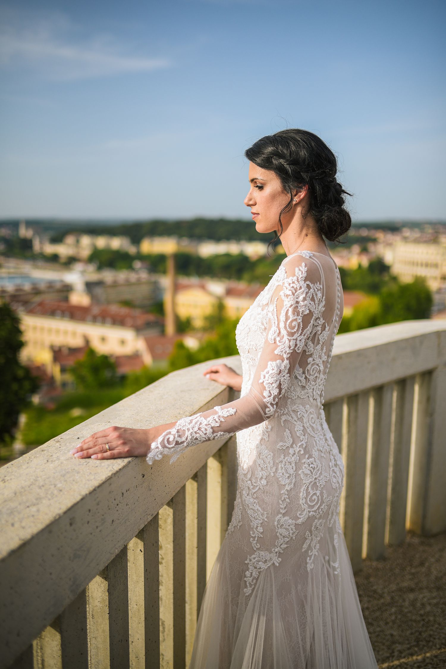 Elegant bride in lace gown standing on a balcony overlooking a scenic European cityscape.